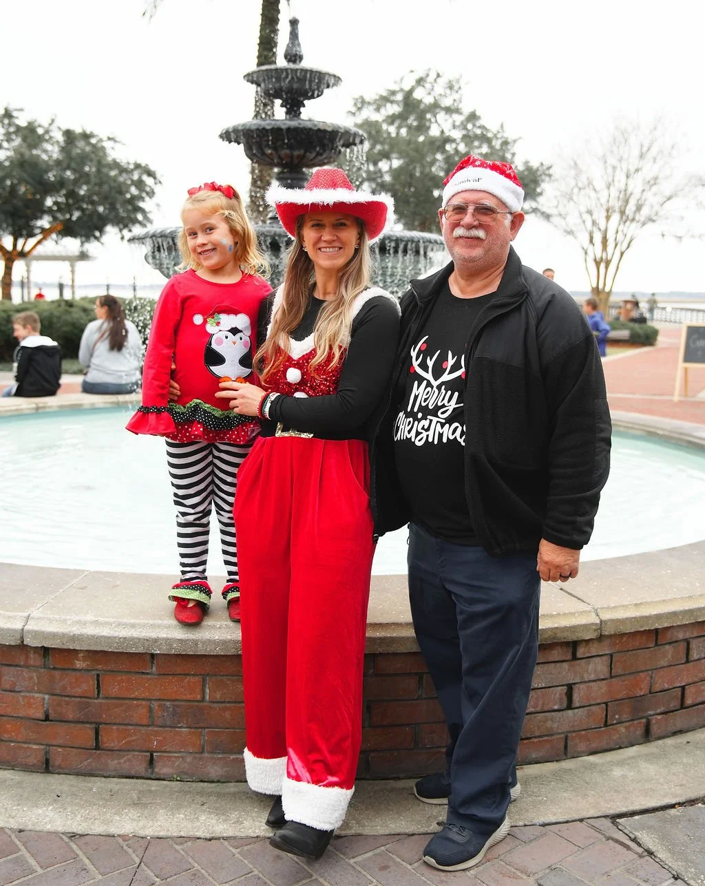 A family dressed in Christmas attire standing in front of a fountain outdoors.