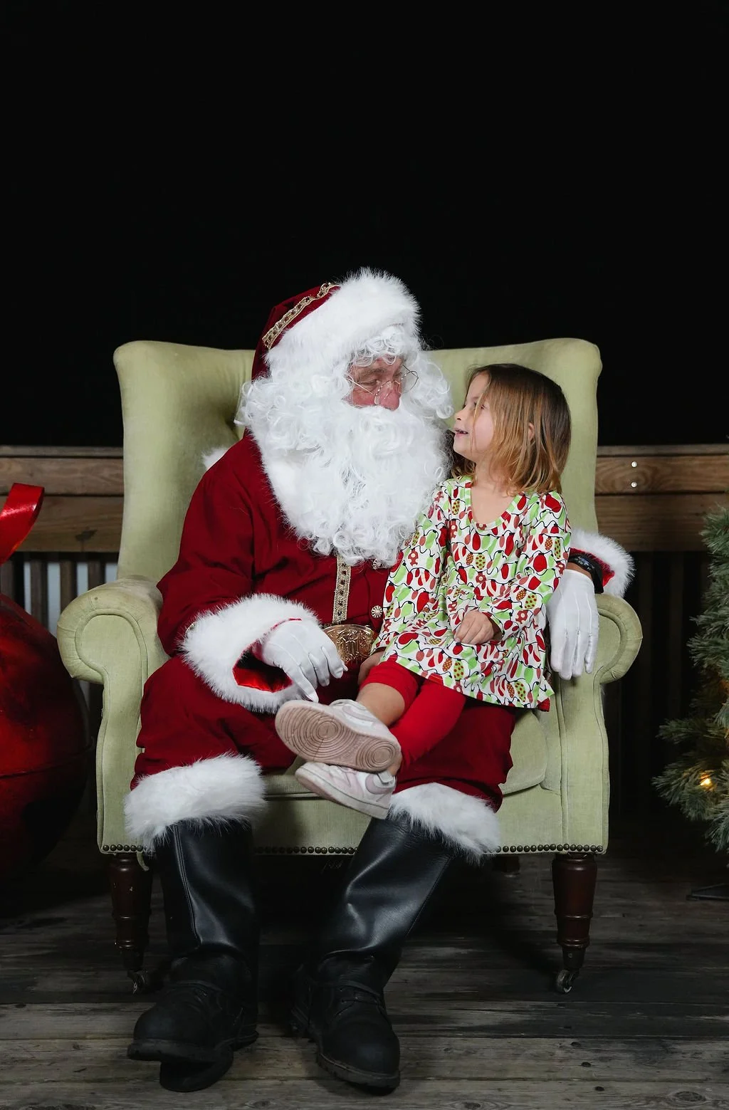 A young girl sitting on Santa Claus's lap during Christmas, indoors with a wooden floor and dark background, Christmas tree partially visible on the right.