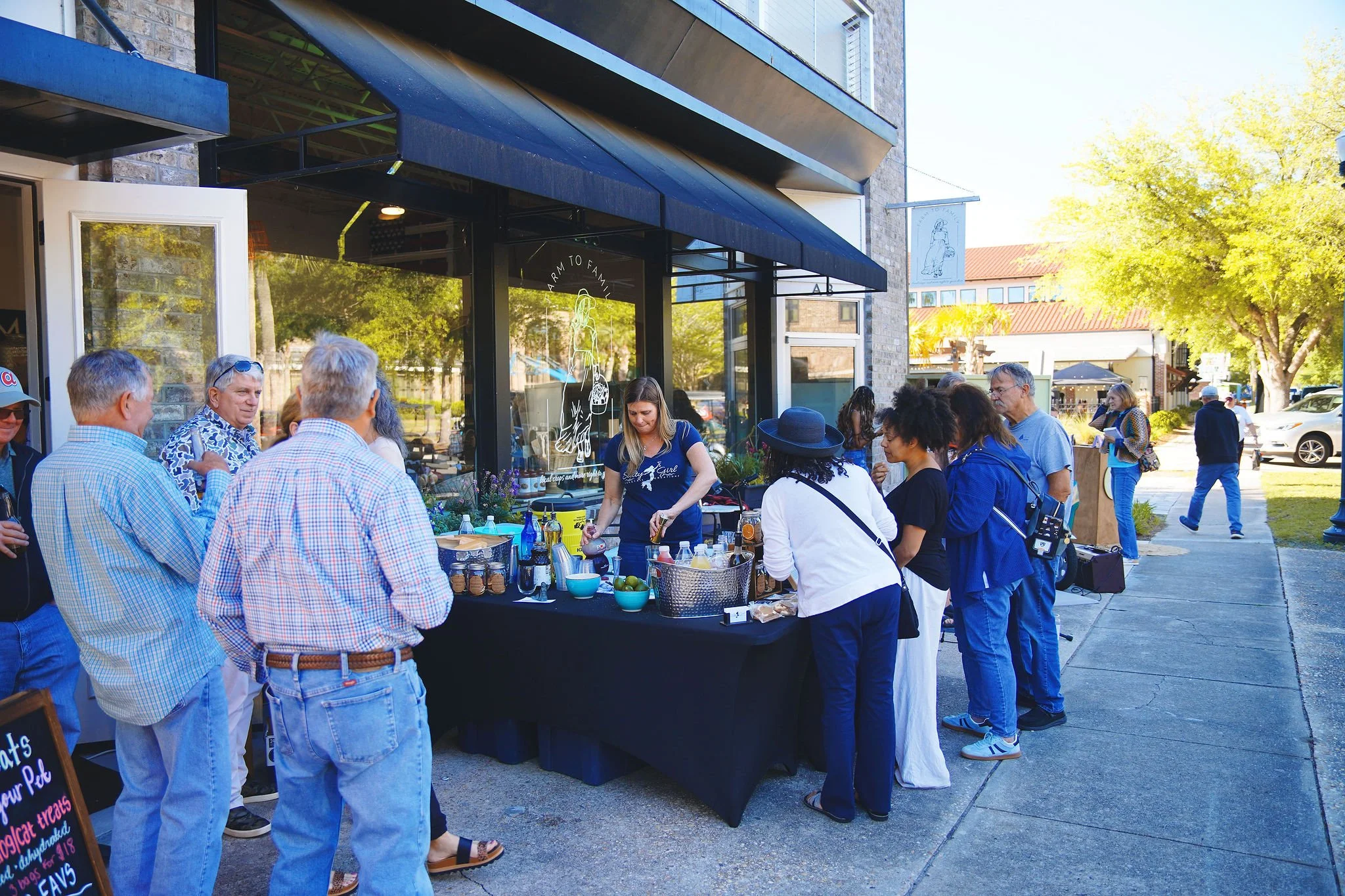 People gather outside a storefront for a coffee tasting event, lining up and chatting on a sunny day in a downtown area with trees and parked cars.