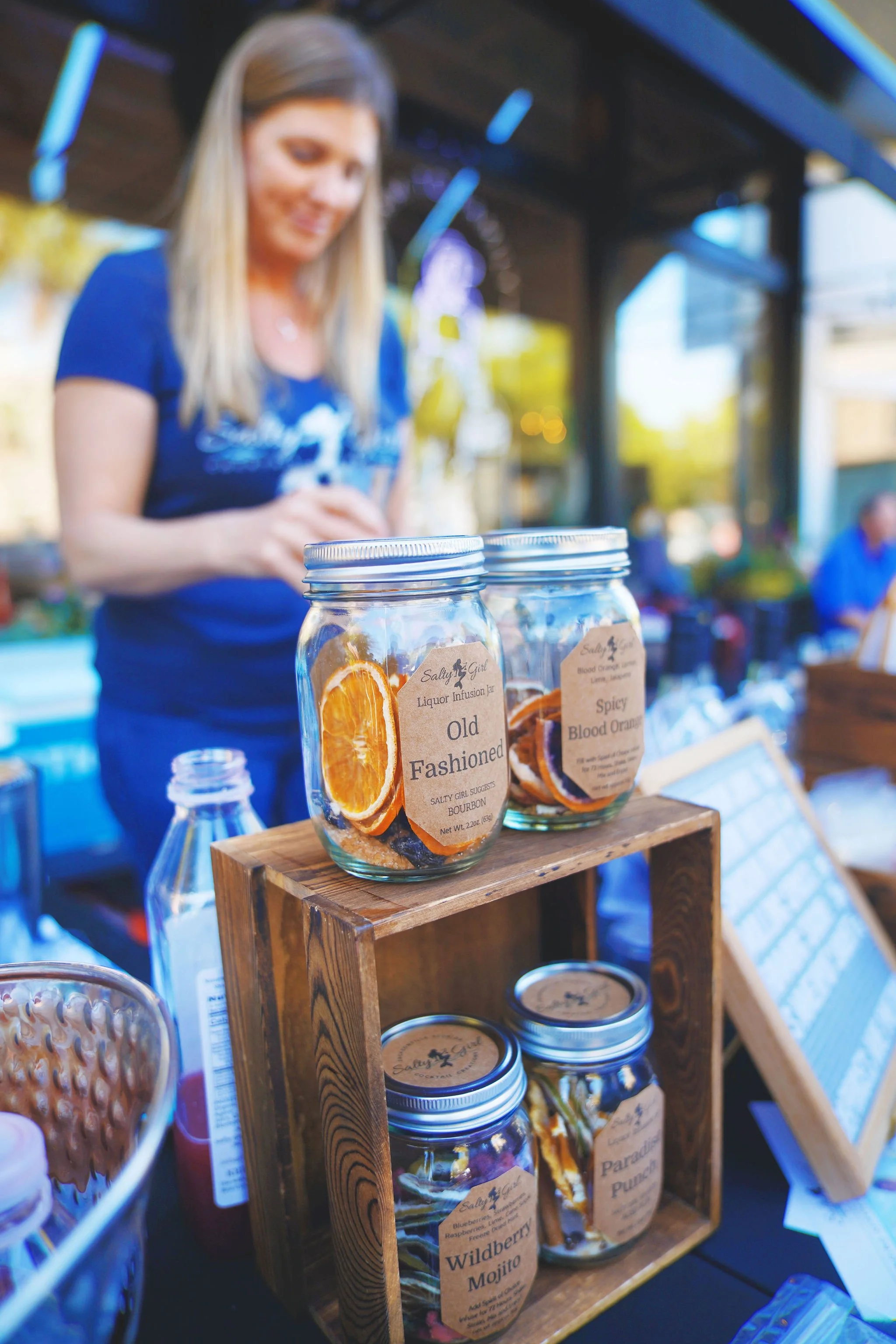 Clear jars with dried fruit slices and labels on display at an outdoor market stall, with a woman in a blue shirt behind them.