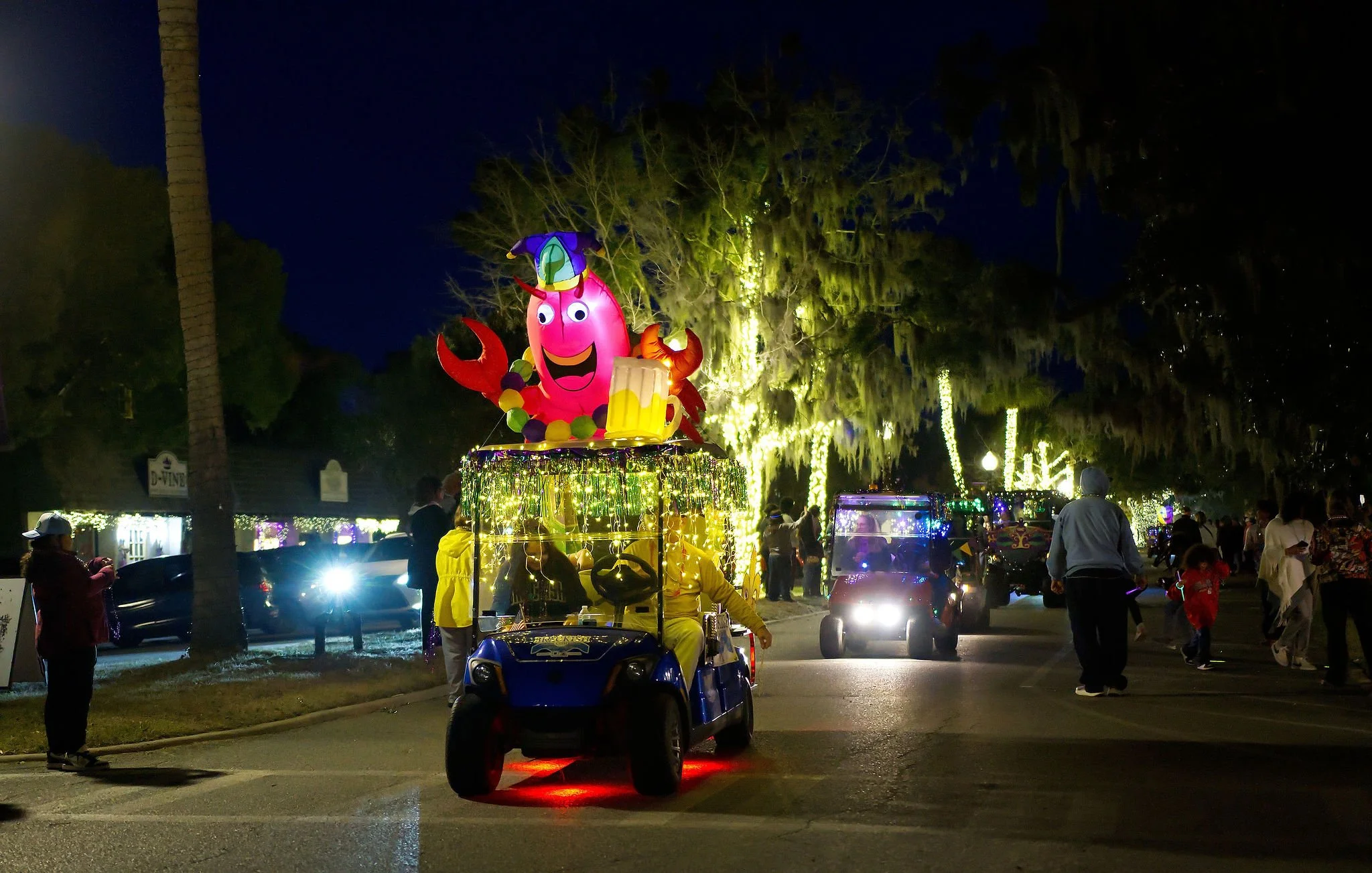 Nighttime parade with decorated golf carts, featuring a large inflatable crab holding a beer mug, surrounded by string lights and people walking along a street lined with illuminated trees.