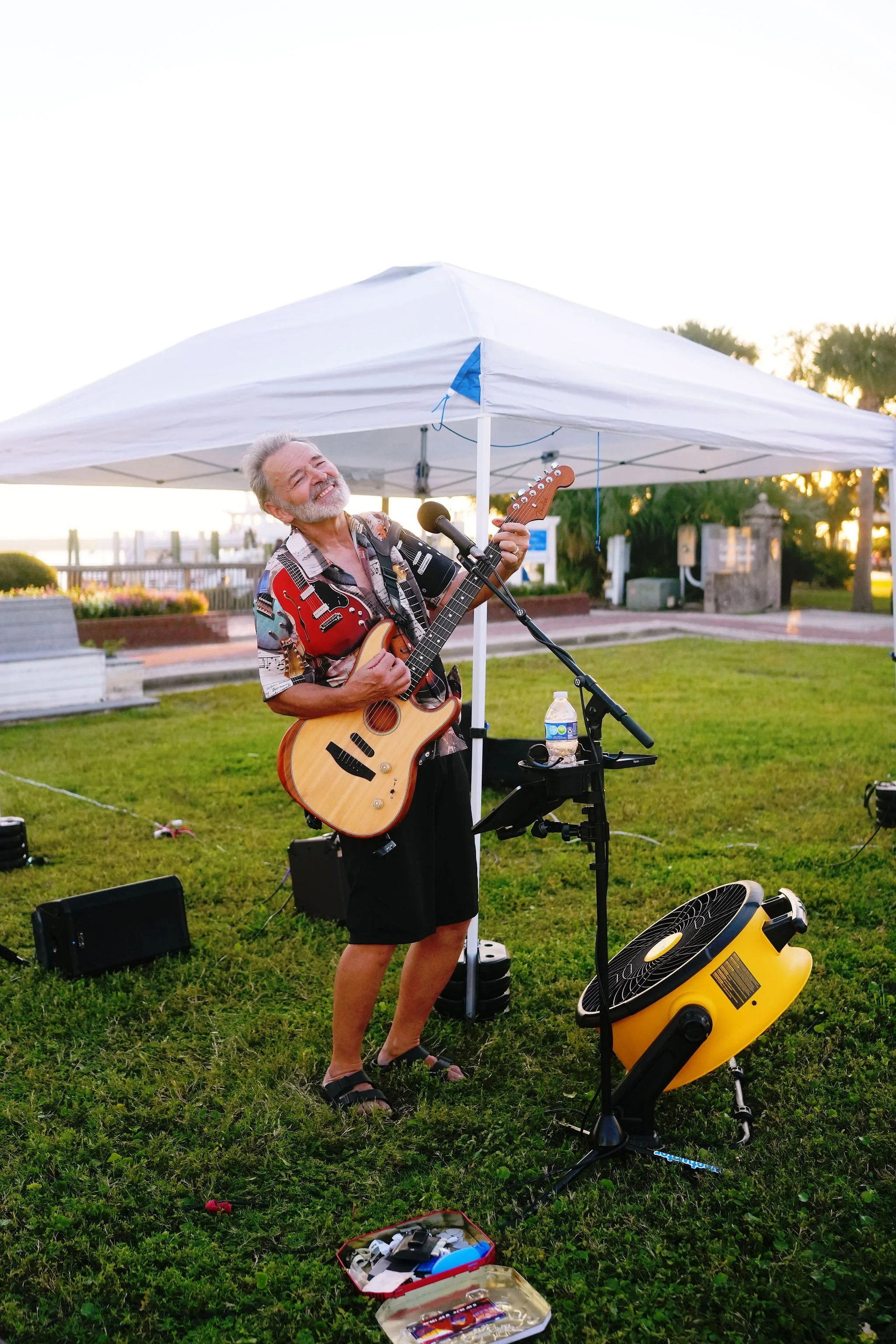 Older man playing an acoustic guitar and singing outdoors under a white canopy tent, with audio equipment and a fan on the grass.