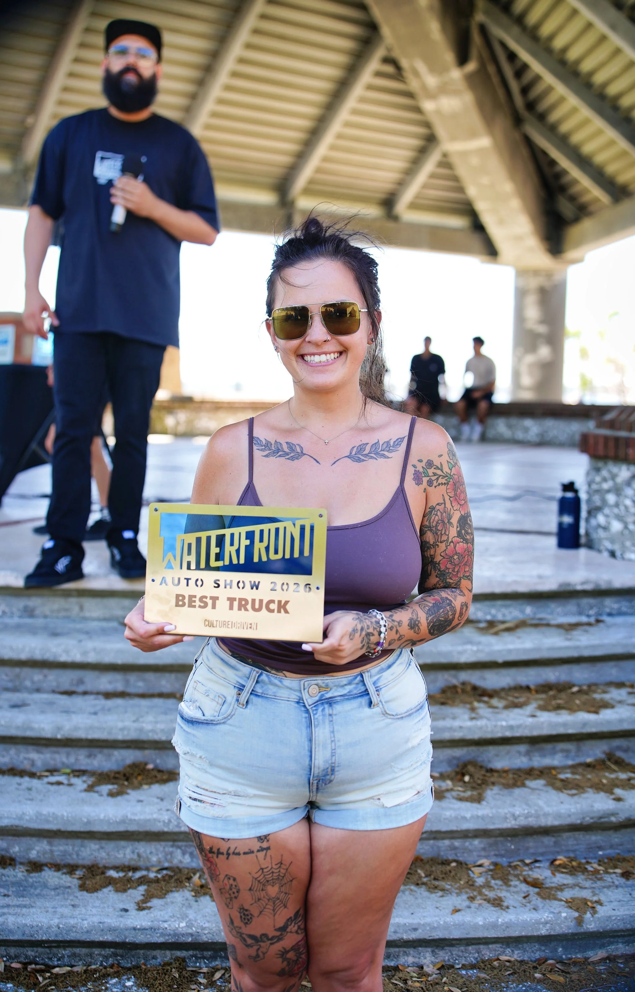 A smiling young woman with tattoos on her arms and leg, wearing sunglasses, a purple tank top, and denim shorts, holding a sign that reads 'Best Truck' at a car show event. In the background, a man with a beard and glasses is speaking into a micropho