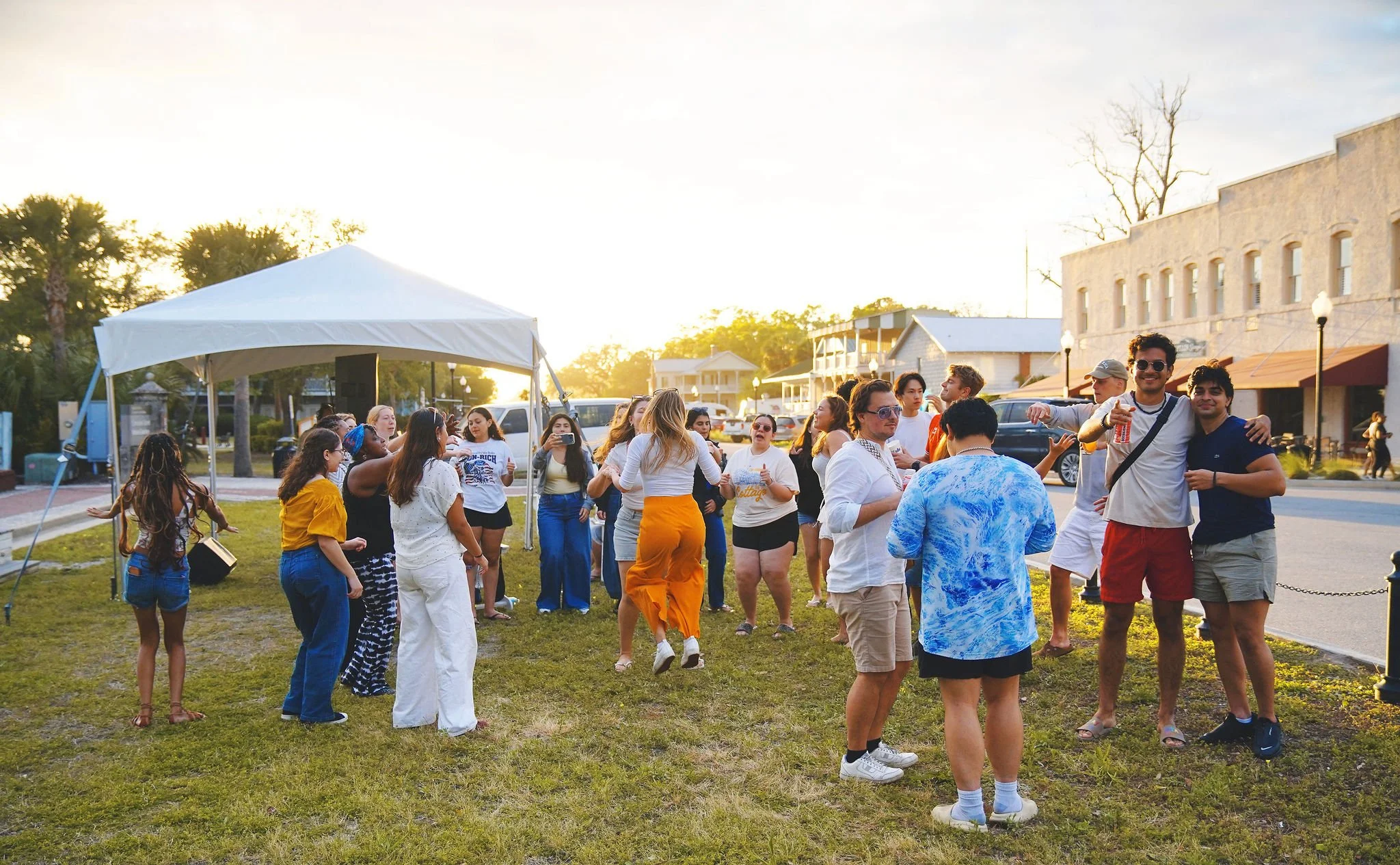 Group of people gathering outside under a tent at sunset, some dancing and talking on a grassy area near buildings and parked cars.