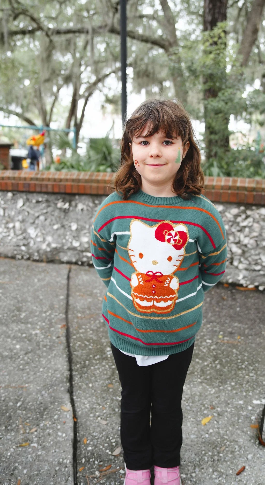 A young girl with brown hair, wearing a festive sweater featuring Hello Kitty, standing outdoors on a gray pavement with trees and a brick wall in the background.
