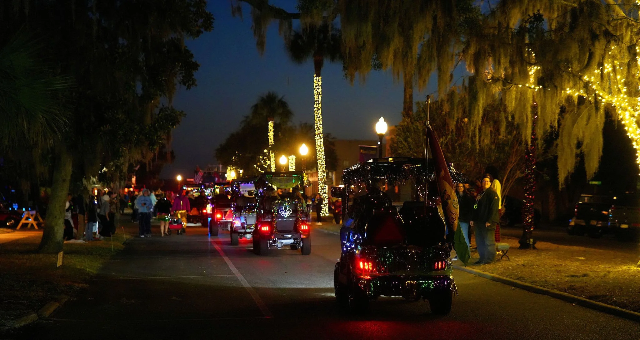 Nighttime scene in a street decorated with Christmas lights, with people gathered and riding golf carts.