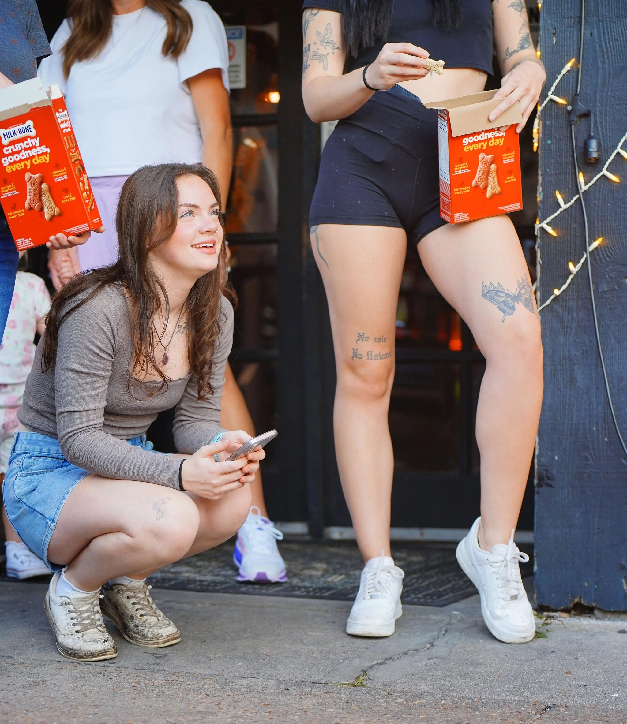 A group of young women, some squatting and some standing, casually hanging out outside. The focus is on their shoes, with a woman in white sneakers, another in white sneakers, and a woman in white sneakers with long socks. One woman is holding a smar