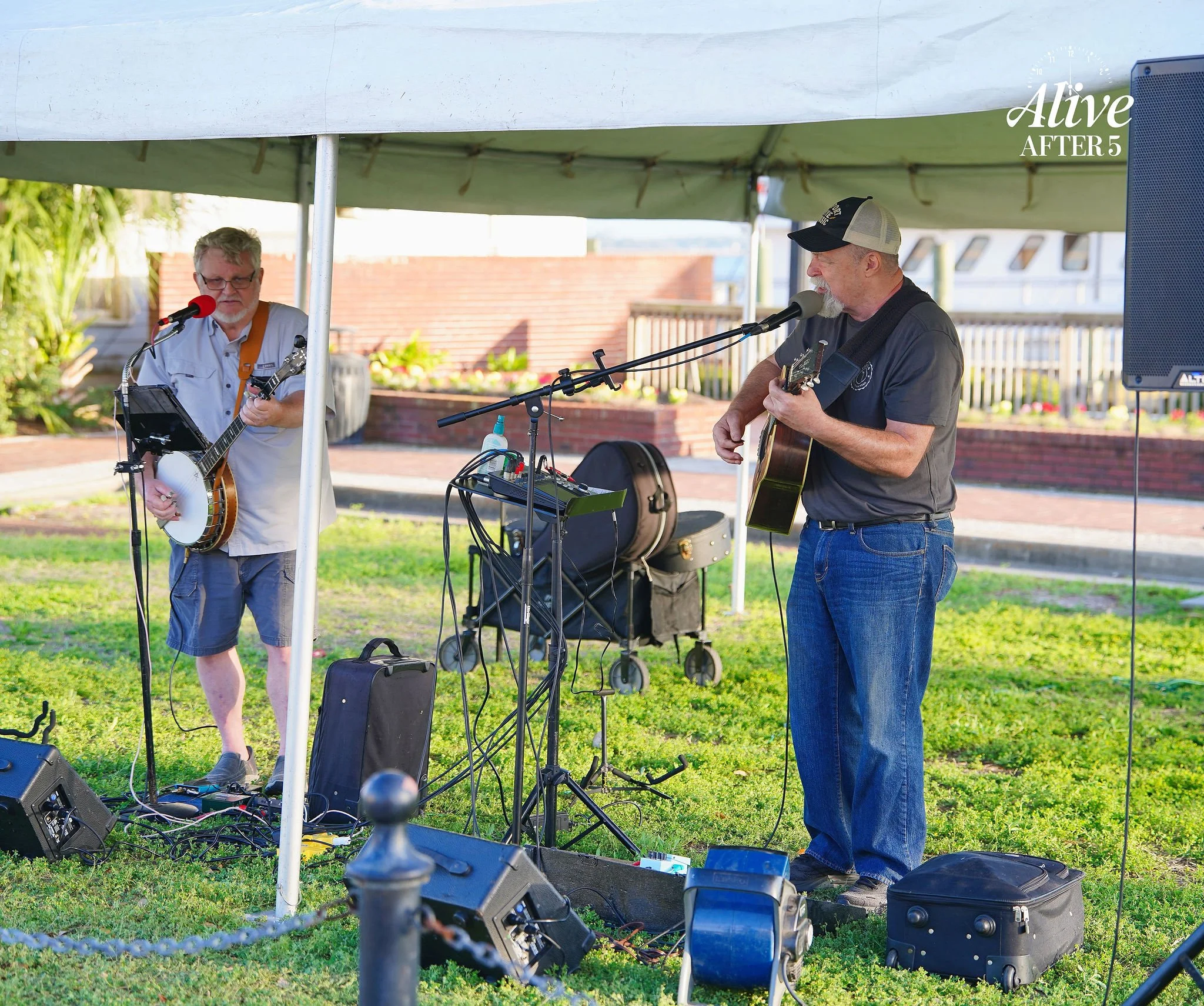 Two men perform music outdoors under a tent, one with a banjo and the other with an acoustic guitar, surrounded by sound equipment and grassy area.