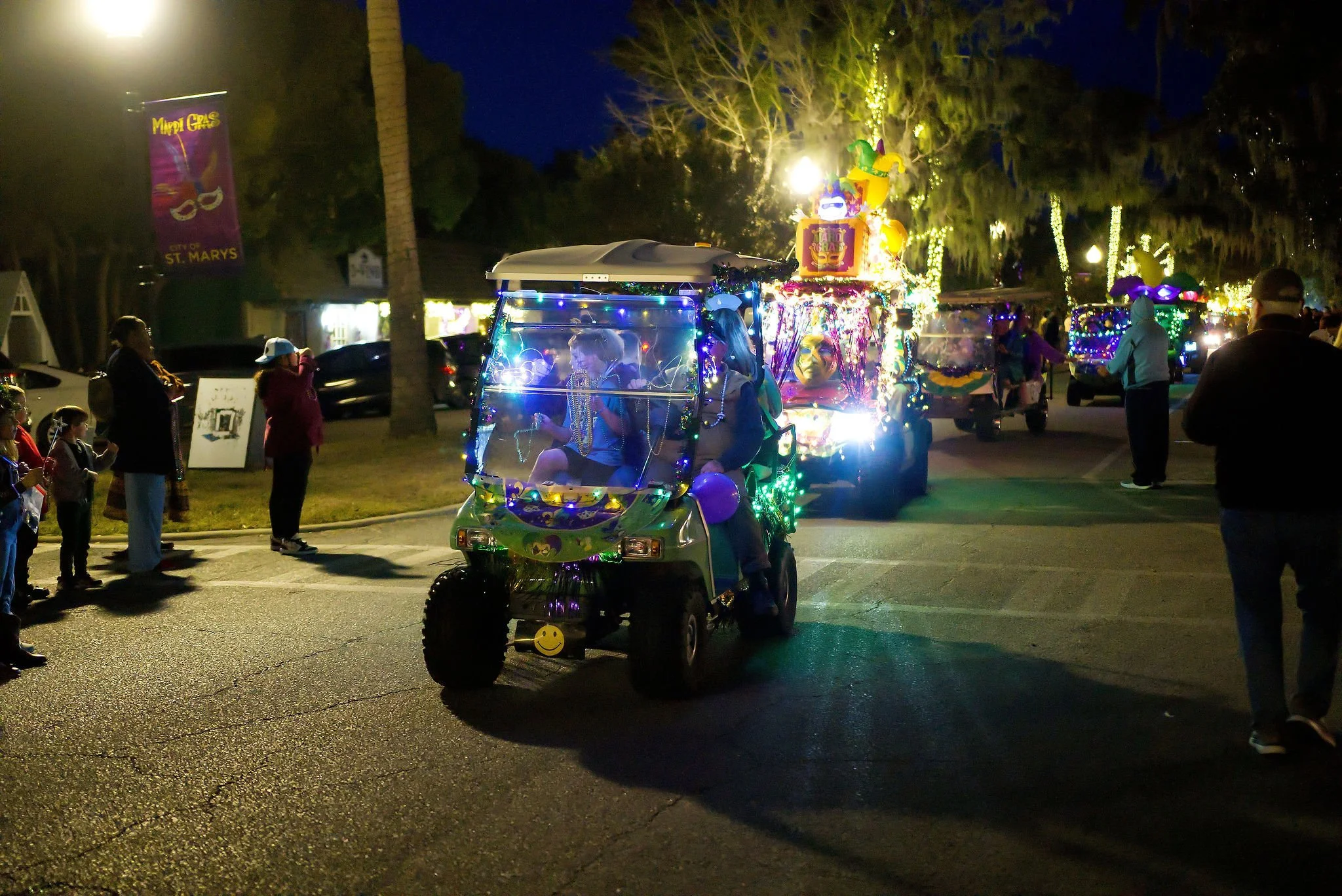 A decorated golf cart with people inside, adorned with colorful lights and decorations, leading a parade at night. Spectators are watching along the street, and other decorated vehicles follow in the background. Trees and buildings are visible under 