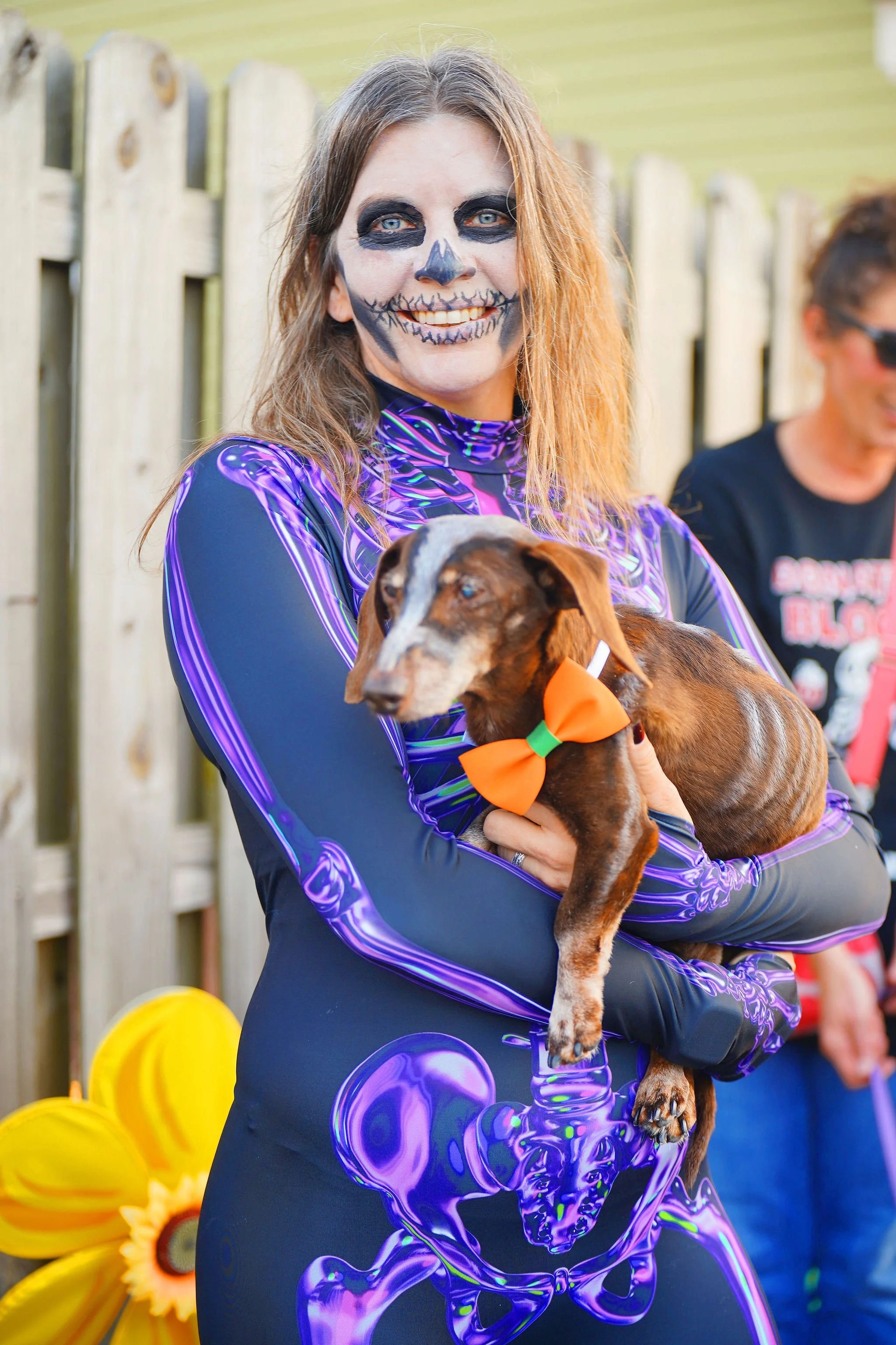 Woman with skeleton face face paint holding a brown dog with a large orange bow around its neck, standing outdoors near a wooden fence.