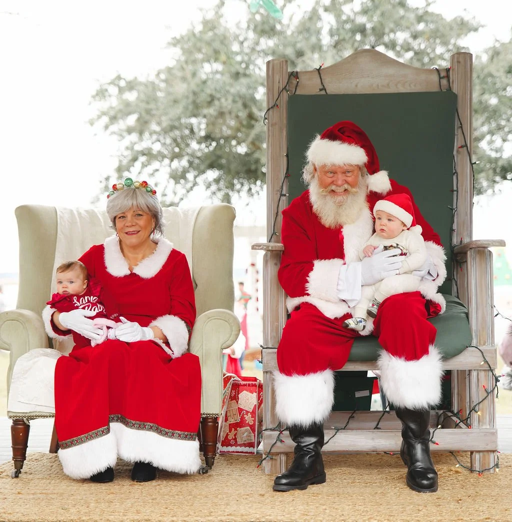 Santa Claus and Mrs. Claus sitting with two children on Christmas-themed chairs indoors, with Christmas decorations and a window showing trees outside.