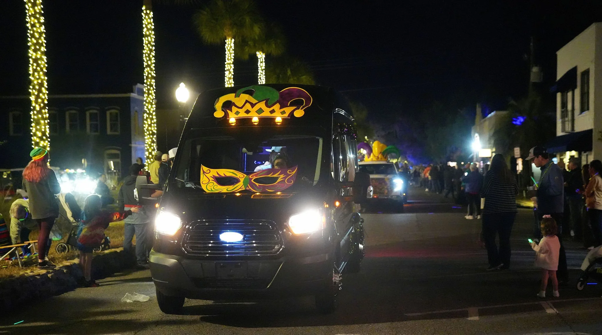 Nighttime parade scene with decorated vehicles and people lining the street, illuminated with string lights on tall trees.