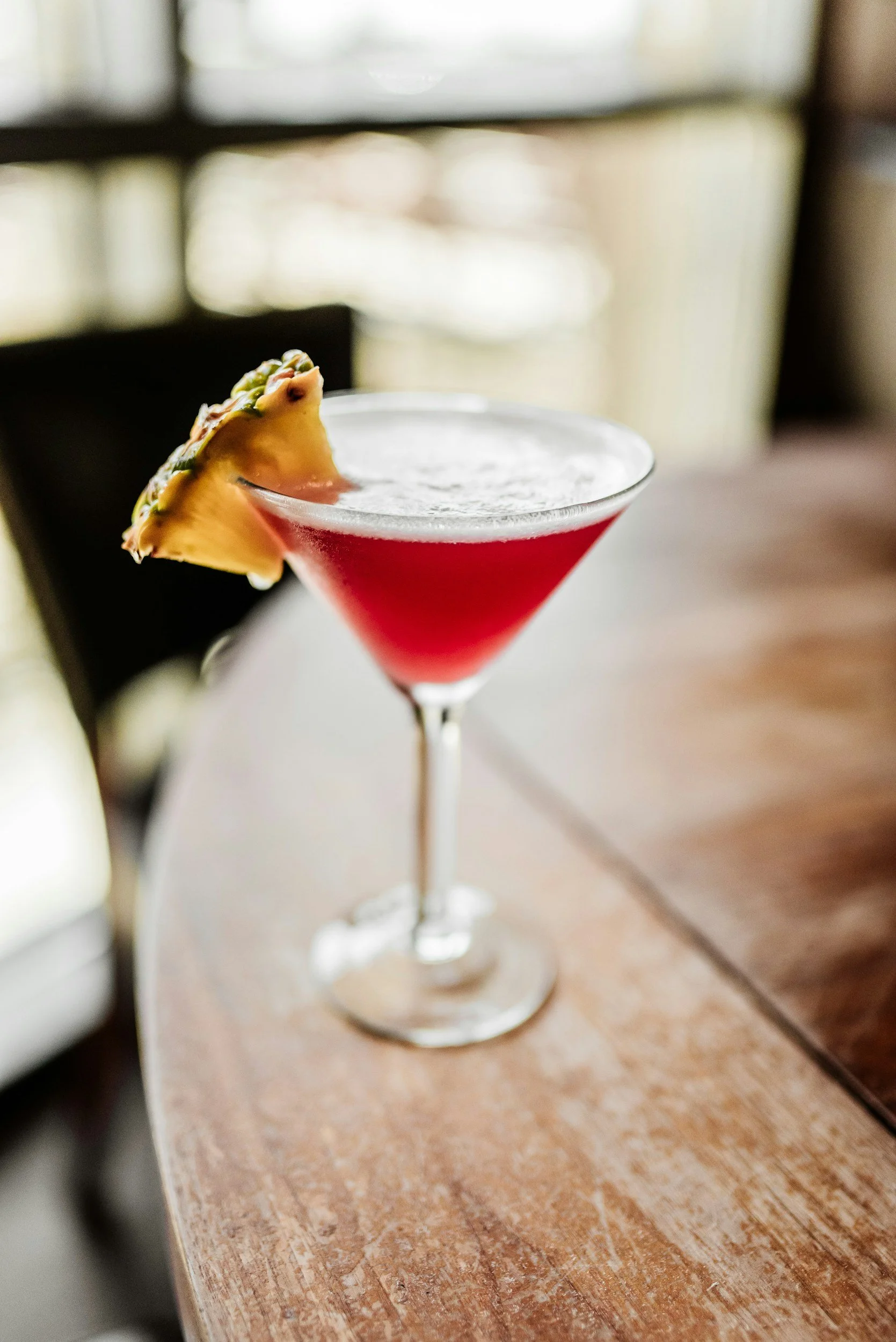 A pink cocktail in a martini glass garnished with a slice of pineapple on a wooden table