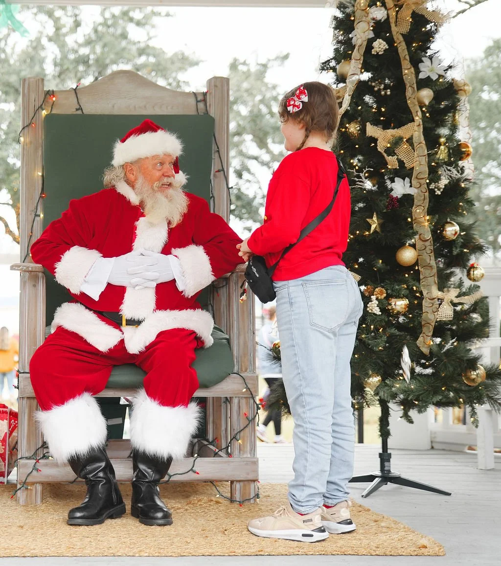 A young girl talking to Santa Claus, who is sitting on a large wooden chair outdoors during Christmas. A decorated Christmas tree stands nearby.
