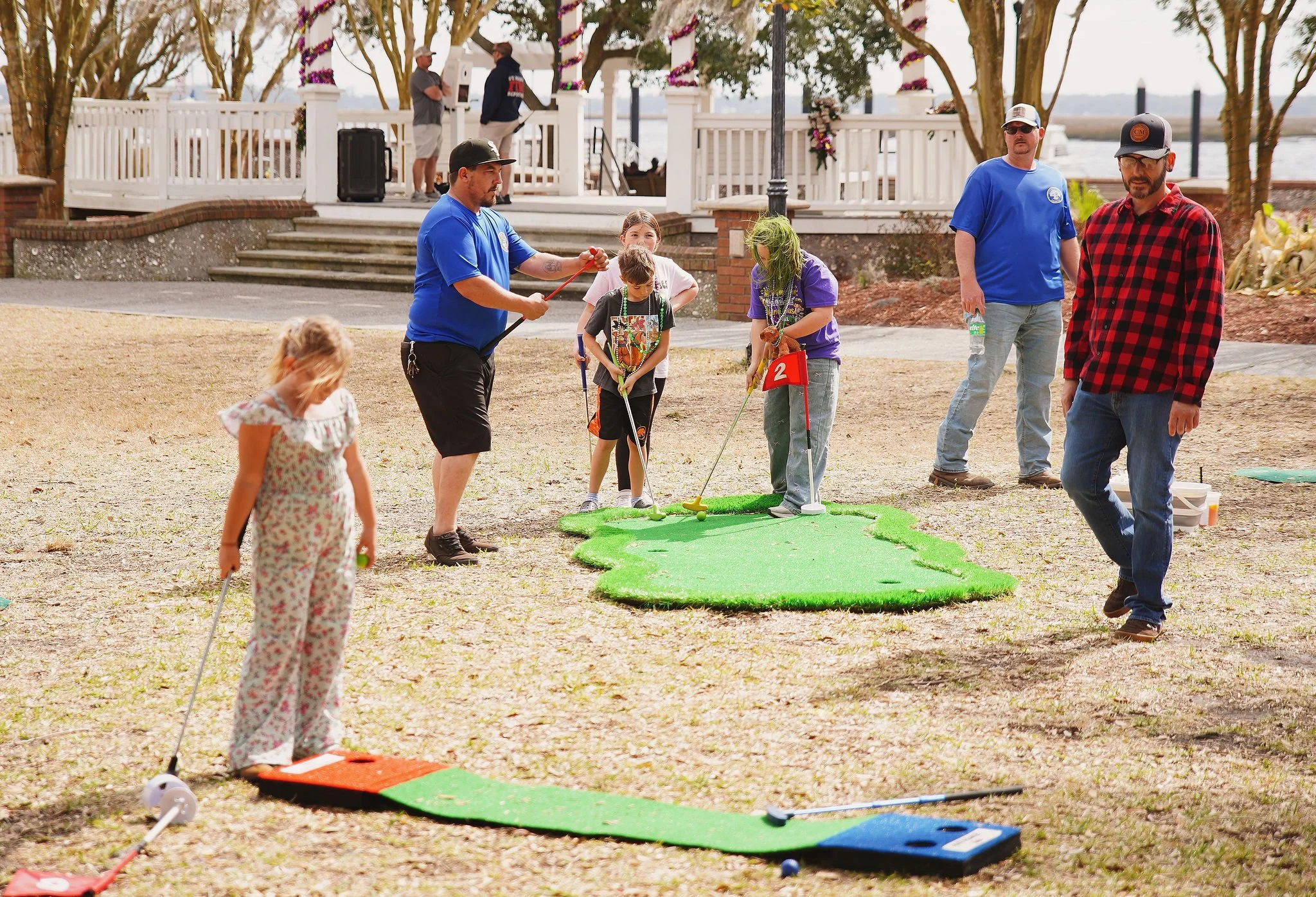 People playing mini golf outdoors on a course with four players, including children and adults, surrounded by trees and a pavilion in the background.