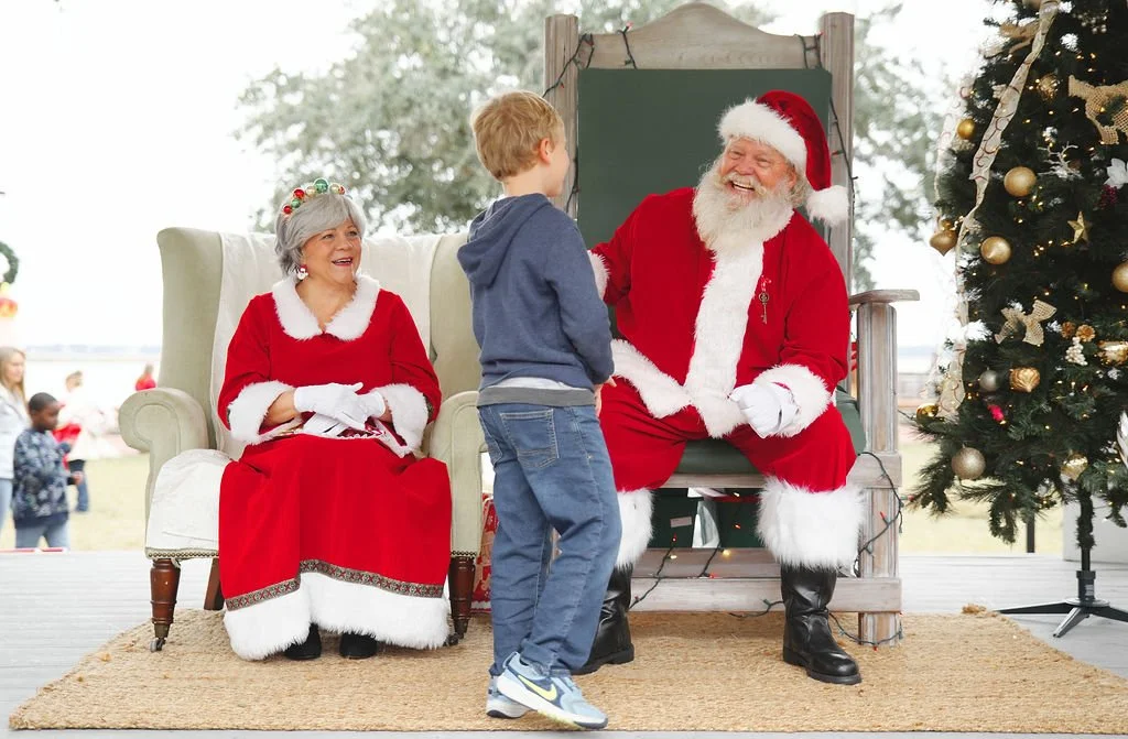 A young boy talking to Santa Claus, who is dressed in a red suit with white trim, seated on a throne. An older woman dressed as Mrs. Claus, in a red dress with white trim and a festive headband, is sitting nearby. They are outdoors with a decorated C