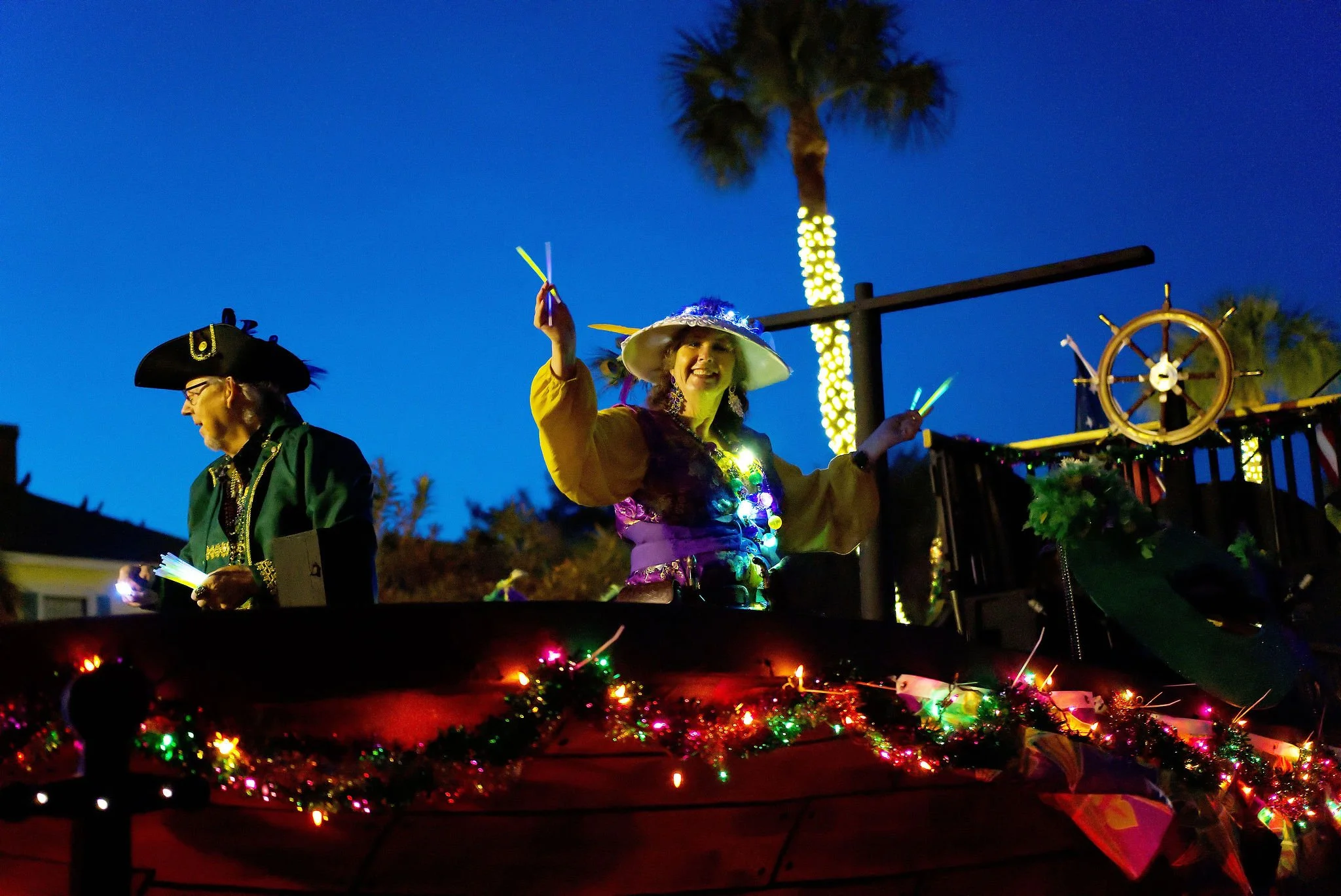 People dressed in colorful costumes, one as a pirate and another as a garden fairy, riding on a decorated parade float with twinkling lights and Christmas greenery, during a nighttime parade with palm trees and a clear blue sky in the background.