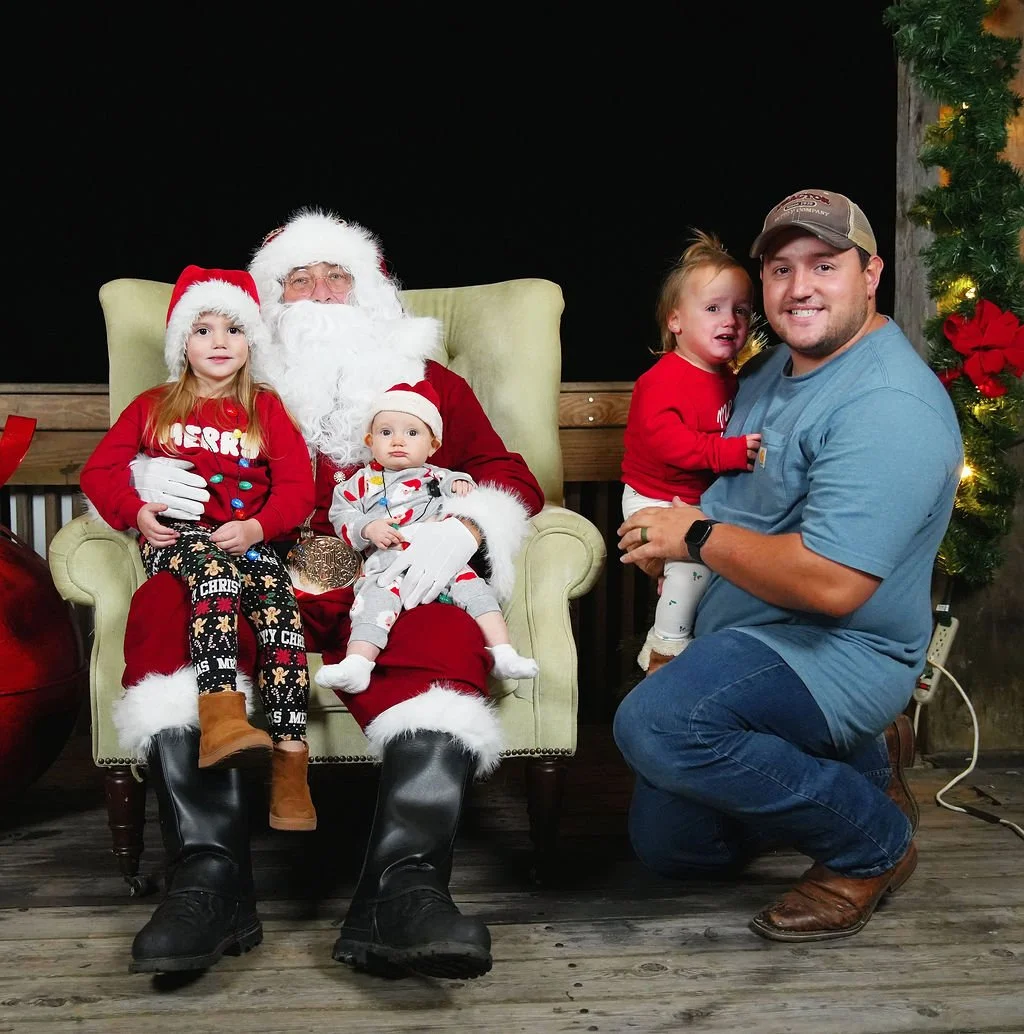 A family taking a photo with Santa Claus during Christmas. Santa is sitting in a green armchair with three children and a man kneeling beside him, holding a young girl. The children are dressed in Christmas-themed clothing, and the man is smiling.