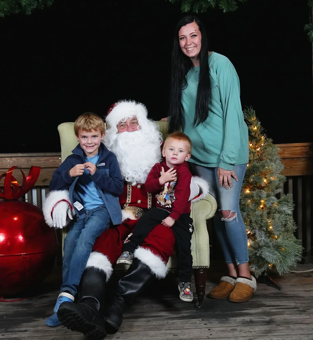 A family with two children taking a photo with Santa Claus at Christmas. The scene includes a decorated Christmas tree, a large red ornament, and festive holiday decorations. The family members are smiling and gathered around Santa, who is seated in 