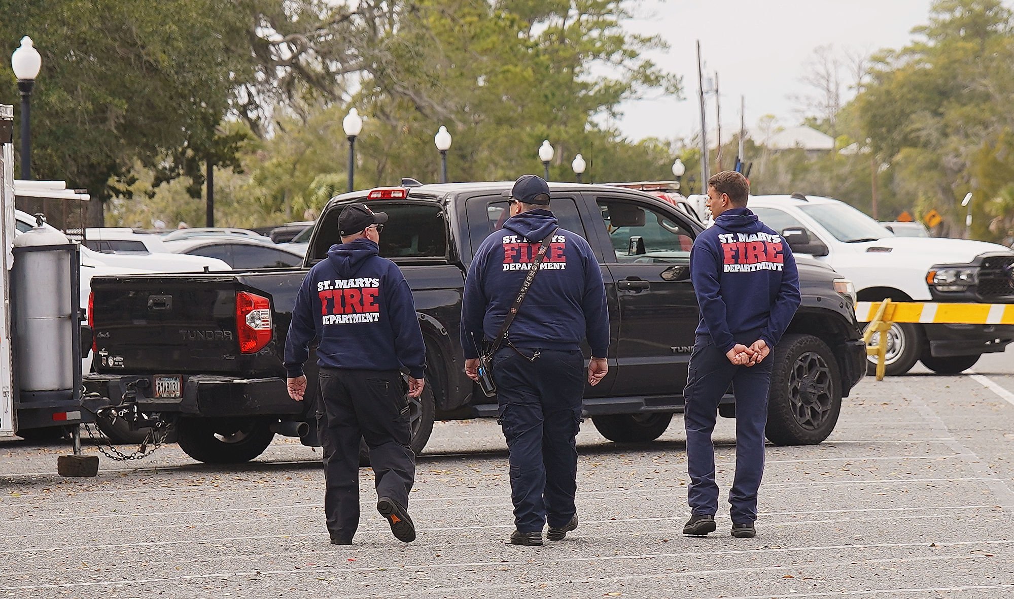 Three firefighters from St. Marys Fire Department walking in a parking lot, with one of them talking to a black pickup truck. The firefighters are wearing navy blue hoodies with the department's name on the back. Several parked cars and trees are vis