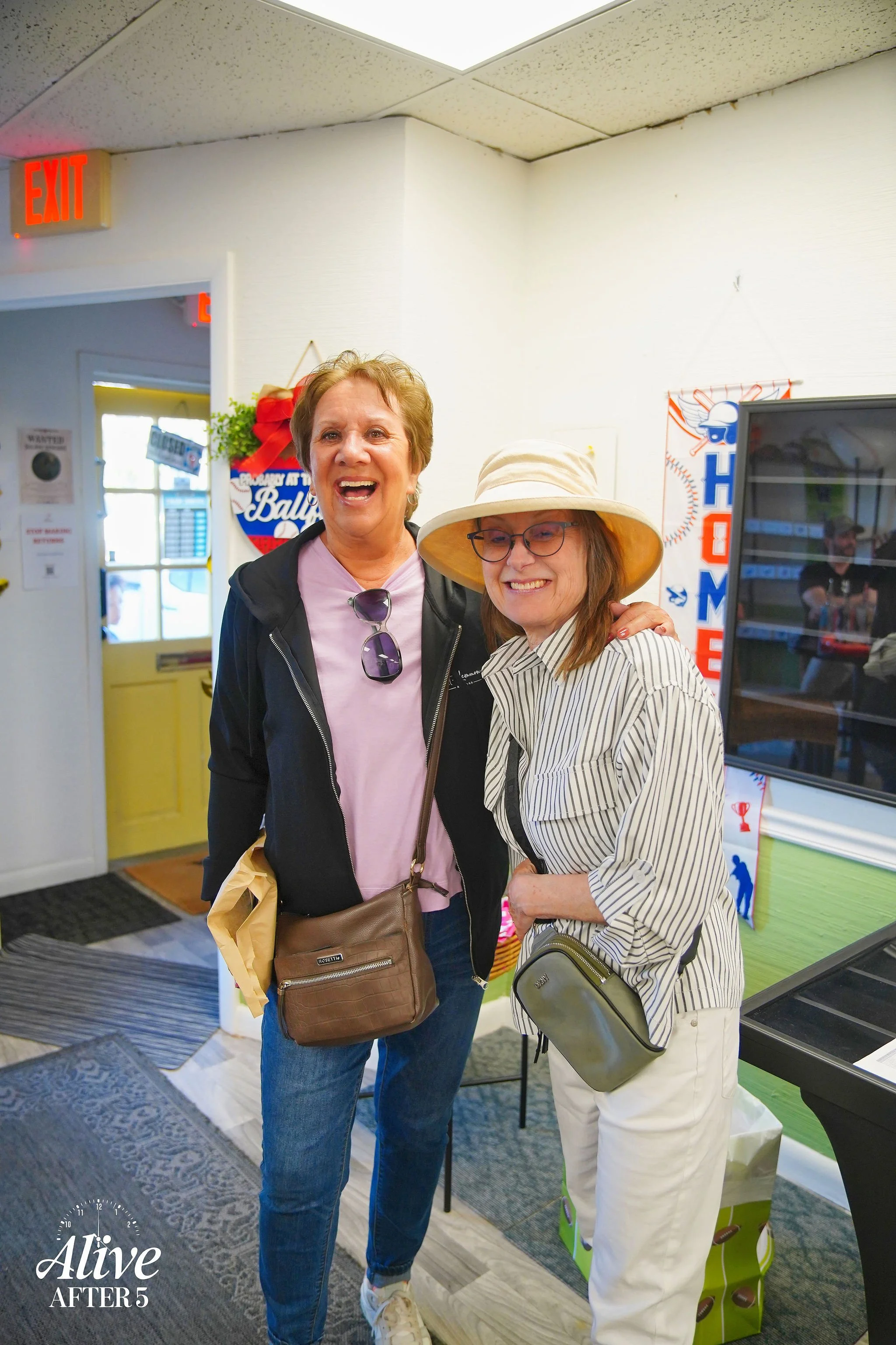 Two women standing together inside a room, smiling and posing for the photo. One woman is wearing a black jacket over a pink shirt with sunglasses hanging from her collar, and the other woman is wearing a wide-brimmed hat, glasses, and a striped shir