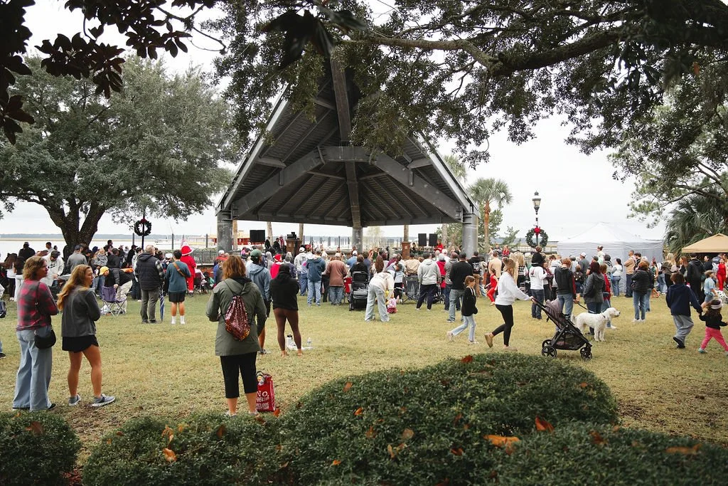 Public outdoor event with a stage, crowd gathered watching performers, holiday decorations, and trees around, during daytime.