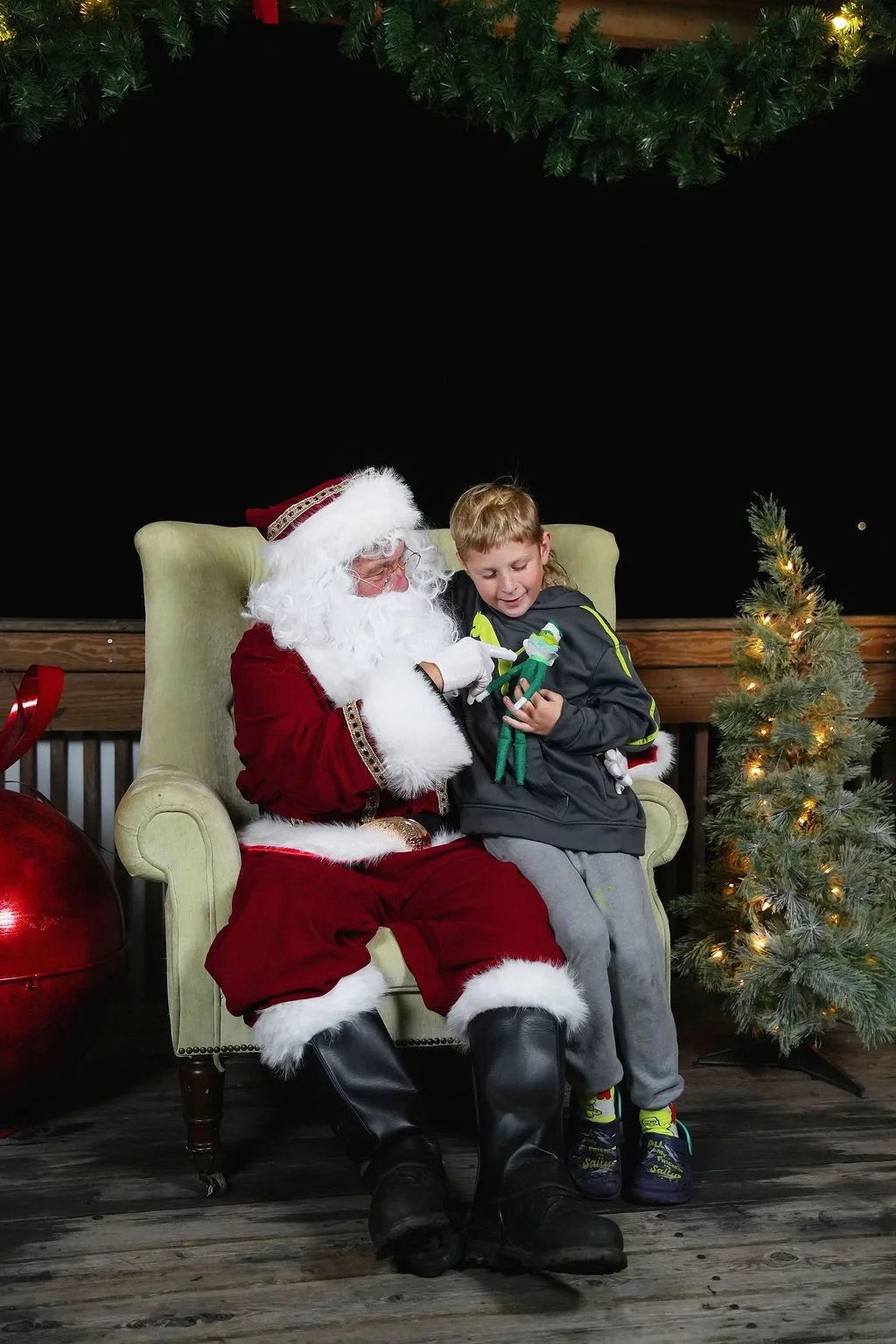 A young boy sitting on Santa Claus's lap, holding a small elf plush toy, in Christmas setting with a decorated small Christmas tree and greenery with lights in the background.
