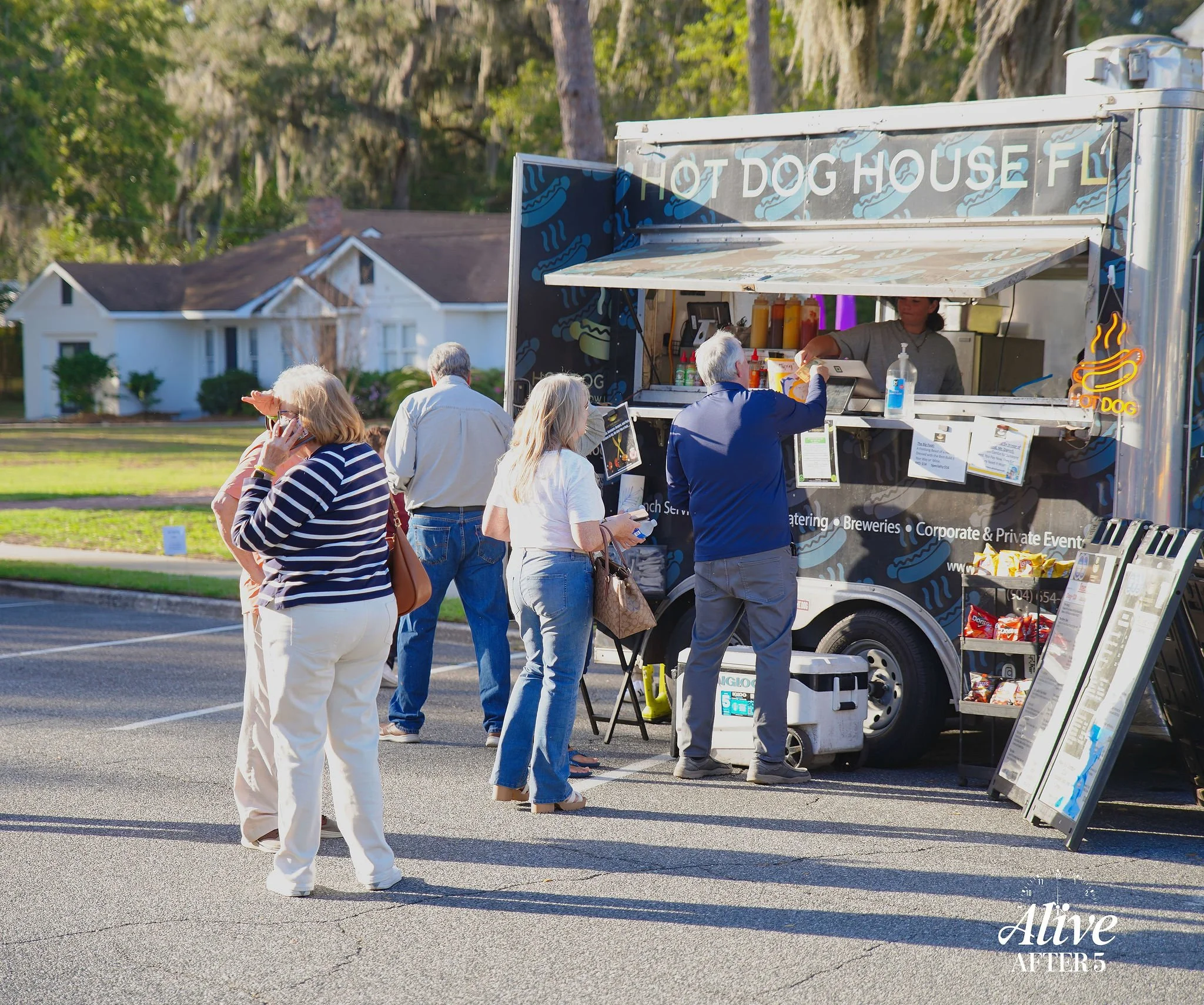 People standing in line at a hot dog food truck in a parking lot, with a house and trees in the background.