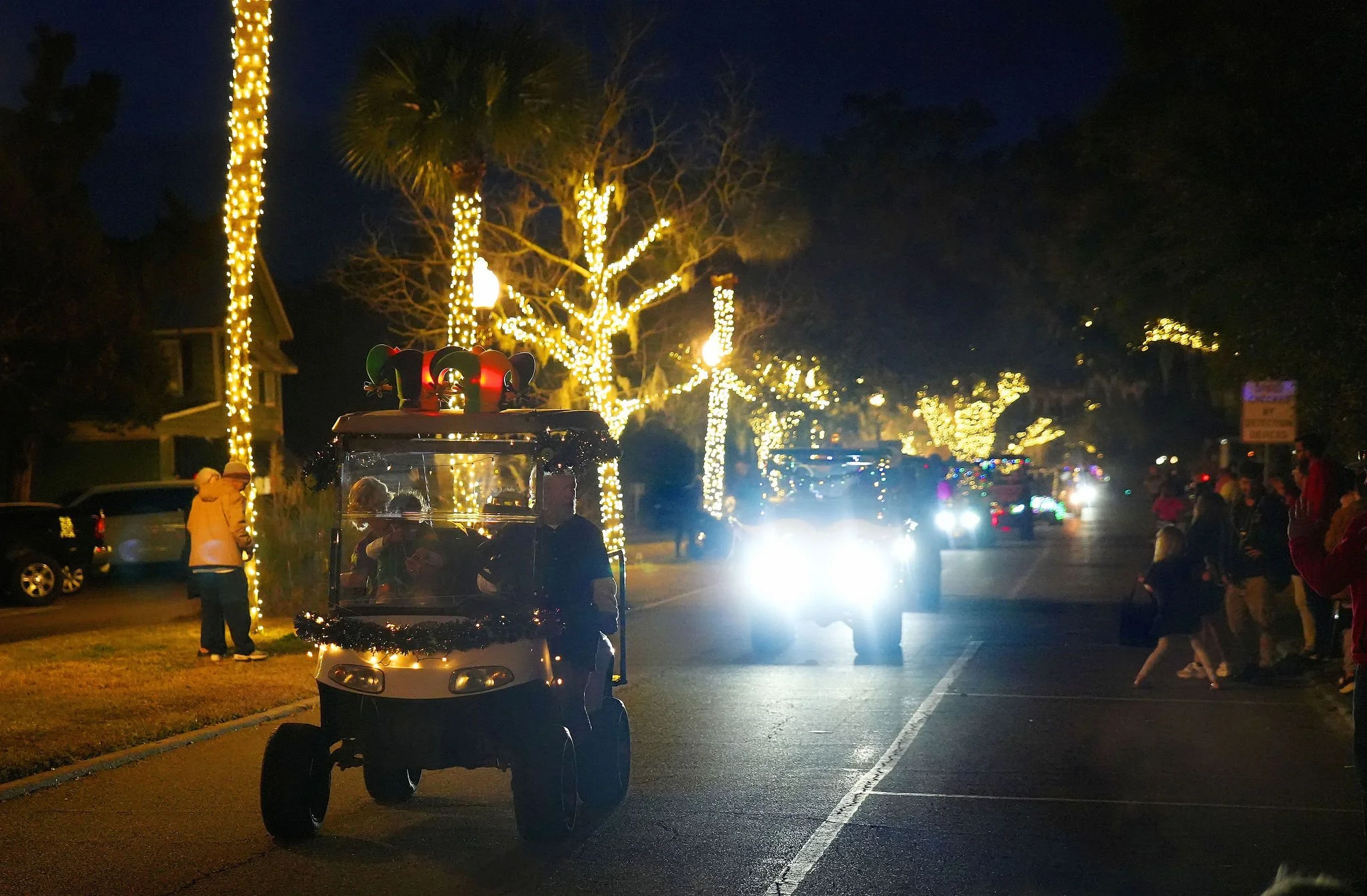 Nighttime parade with a decorated golf cart in the foreground and people on the sidewalk, illuminated trees with string lights, and passing vehicles with bright headlights.