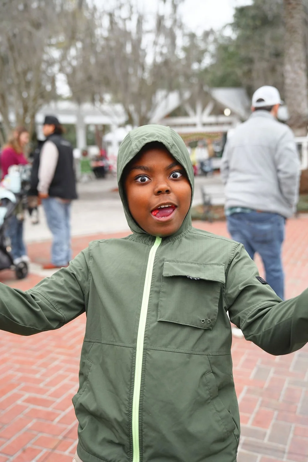 A young boy wearing a green hooded jacket with a surprised and playful expression, sticking out his tongue, in an outdoor setting with people walking and trees in the background.