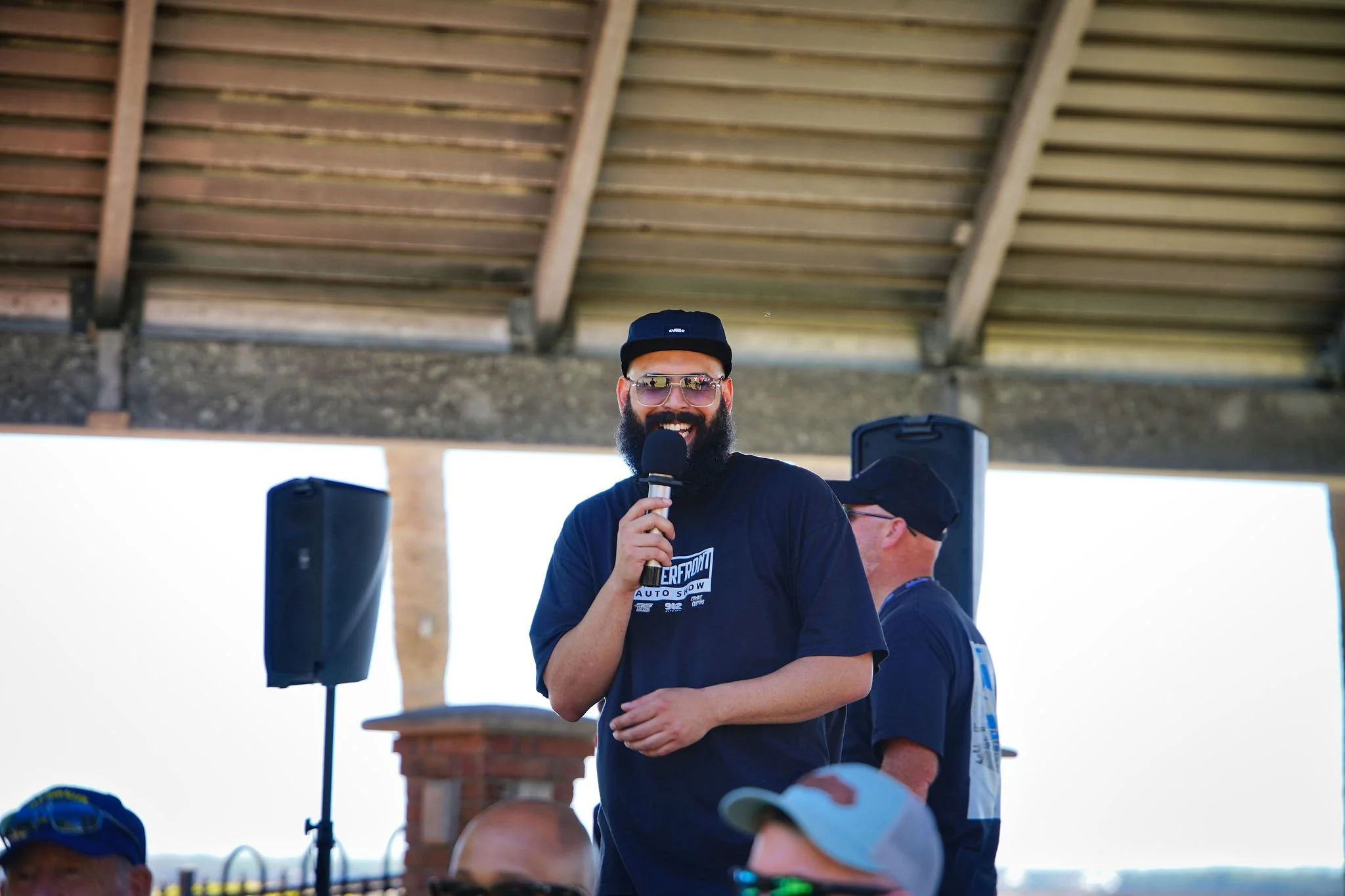 A man with a beard and sunglasses smiling while speaking into a microphone at an outdoor event under a wooden shelter.