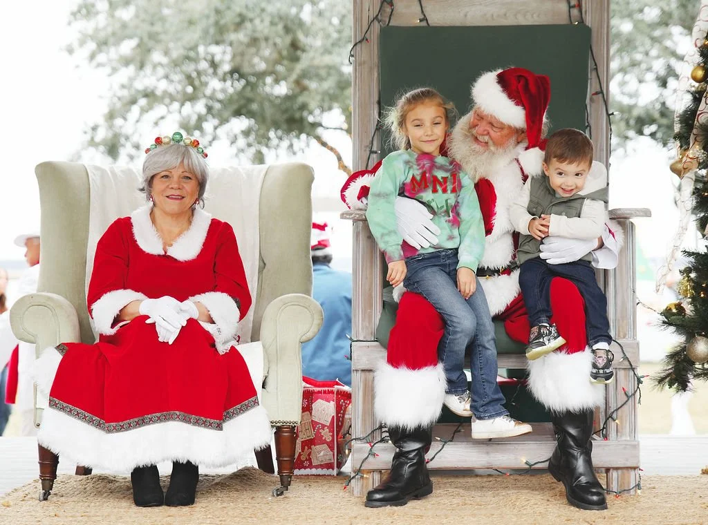 A woman dressed as Mrs. Claus sitting on a chair and two children sitting on Santa Claus's lap, who is wearing a Santa suit, during a Christmas celebration.