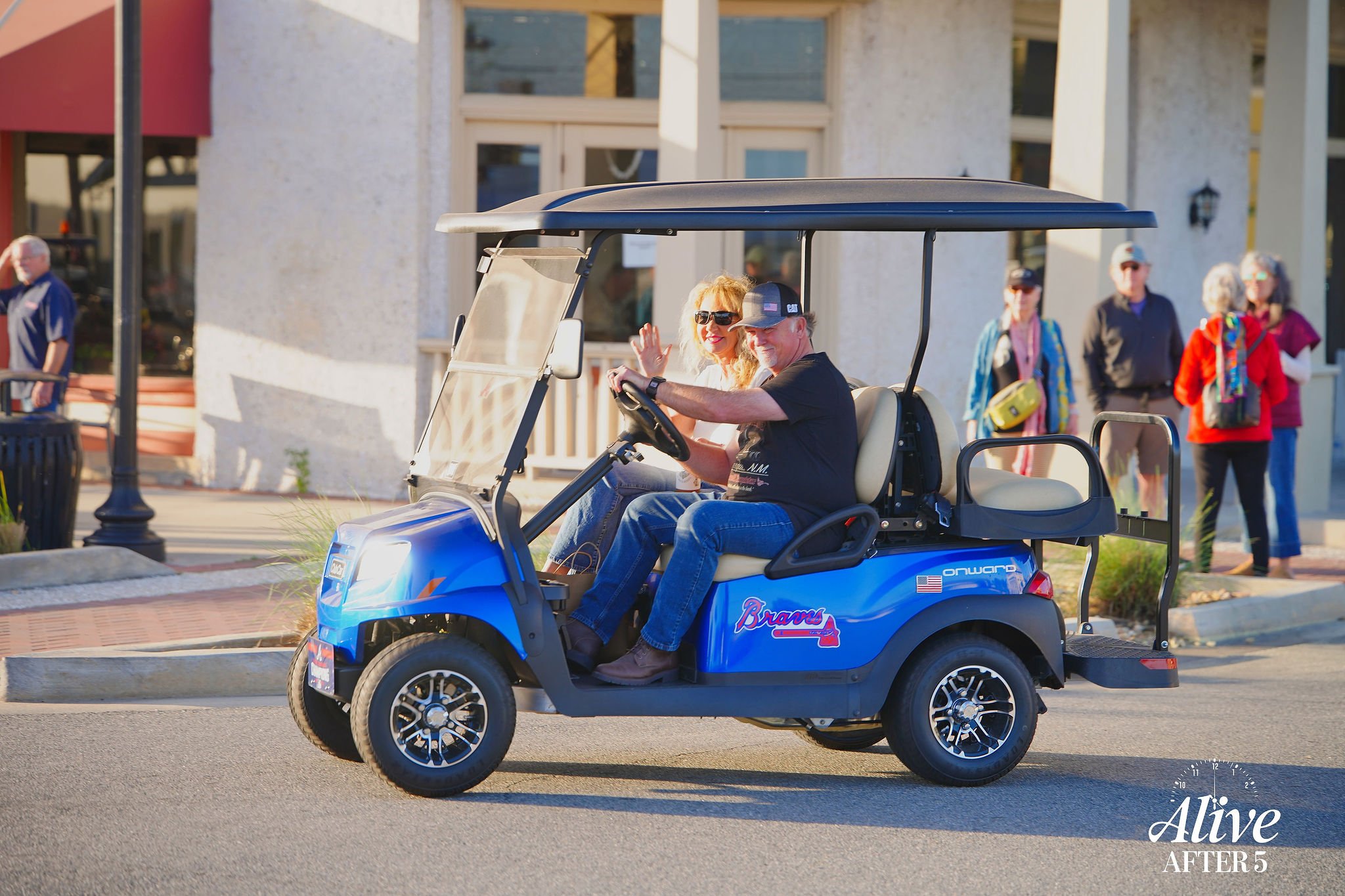 A man and a woman riding in a blue golf cart on a city street. The man is driving, wearing a black shirt, jeans, and a cap. The woman is sitting beside him, wearing sunglasses, a white top, and blonde hair, waving. In the background, there are severa