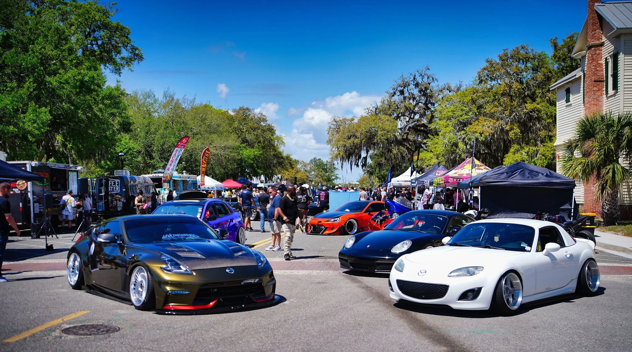 Car show featuring modified sports cars parked on a city street with people walking around, tents, and vendor booths in the background. There are various models of cars, including a black sporty car, a white convertible, and a blue vehicle, against a