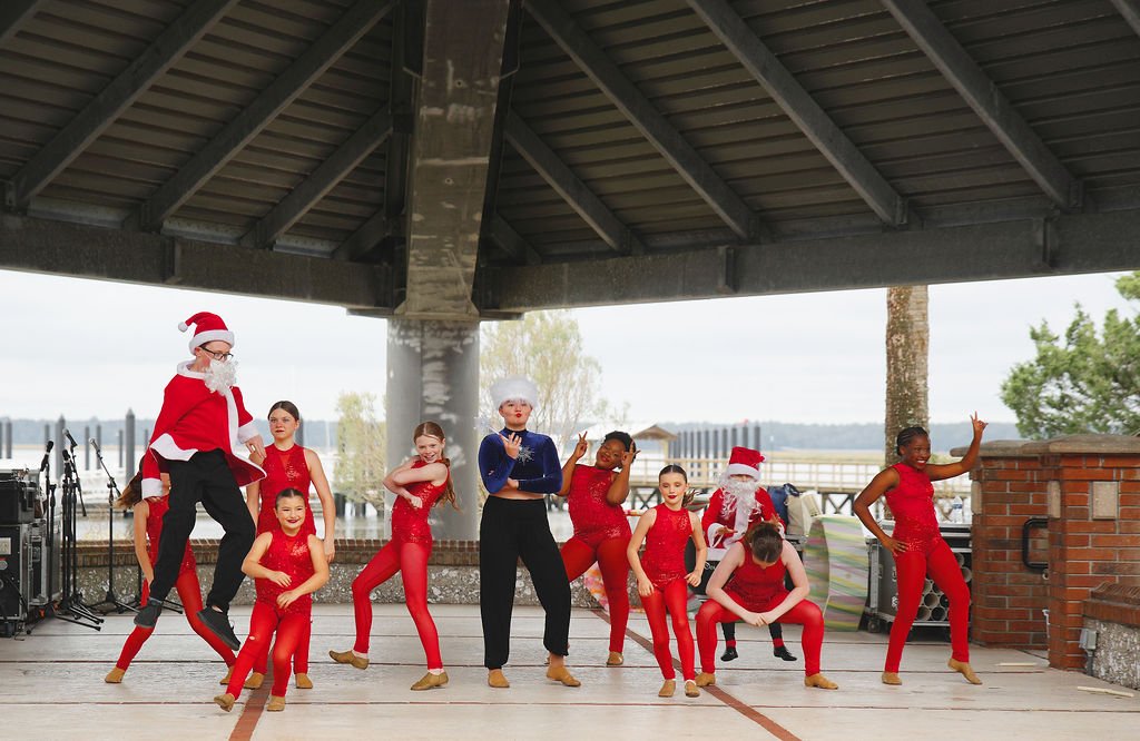Children and adults dressed in festive holiday costumes, including Santa Claus, performing a dance on an outdoor stage under a large shelter with lake and trees in the background.