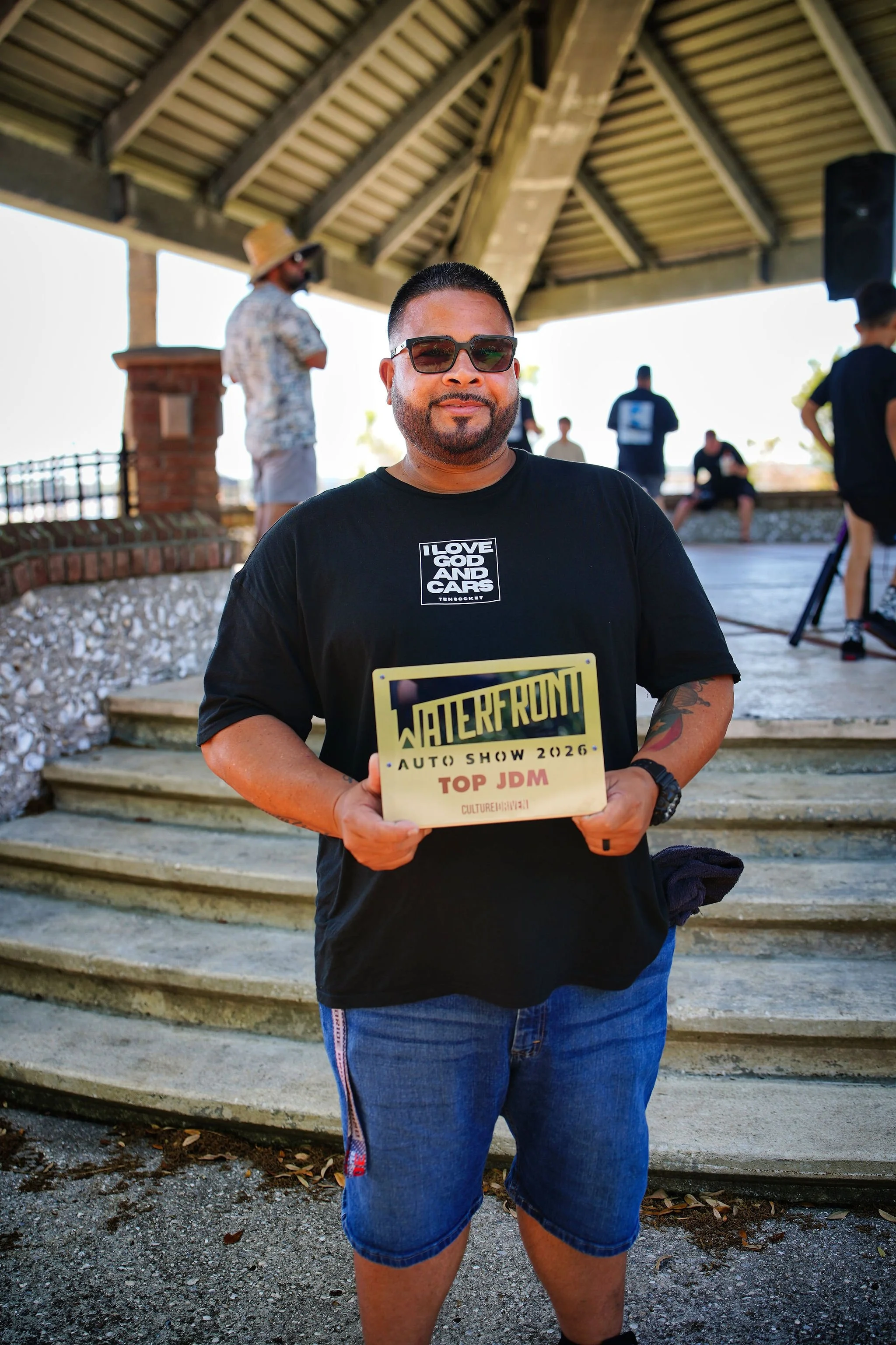 Man holding an award plaque at Waterford Auto Show 2026, Top JDM category, outdoors under a pavilion with other people in the background.
