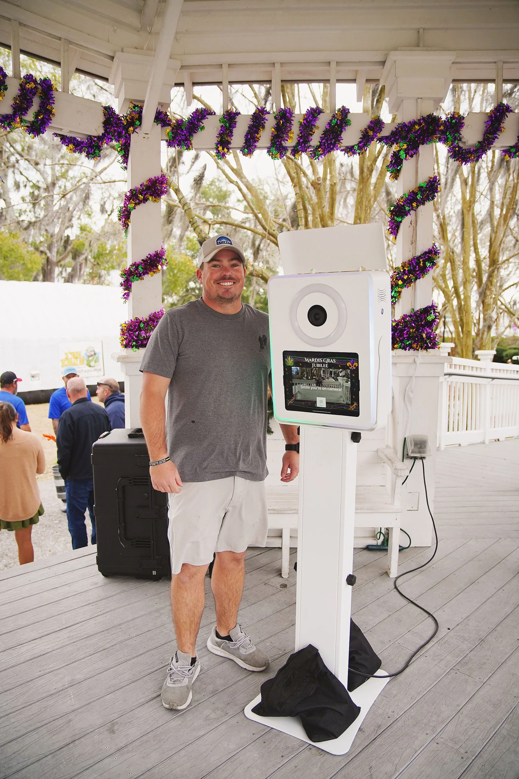 A man smiling and standing next to a photo booth at a Mardi Gras celebration, decorated with purple, green, and gold beads, in an outdoor pavilion with trees in the background.