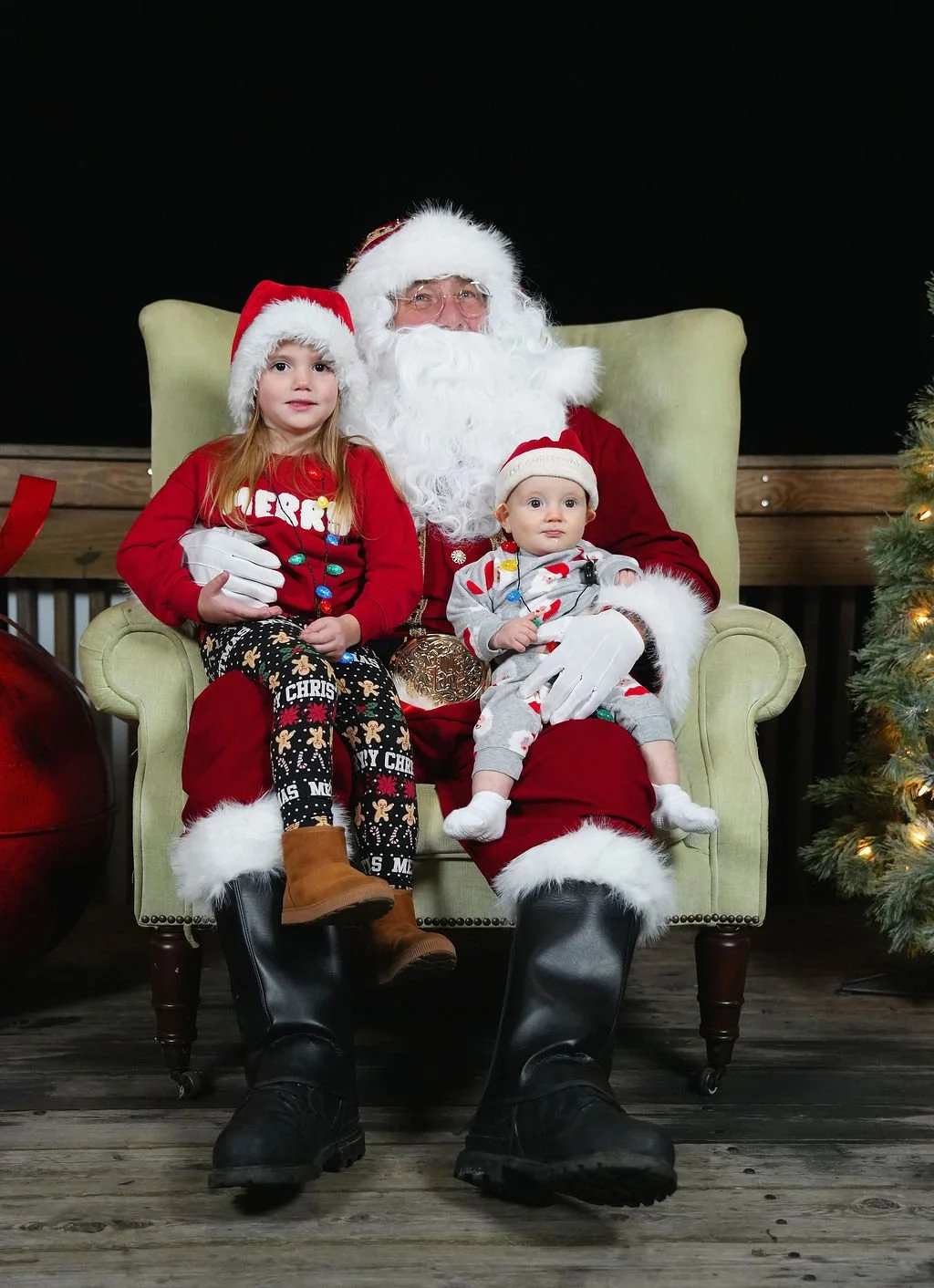 Santa Claus sitting in a green armchair with two young children, a girl and a boy, on his lap during Christmas. The children are dressed in festive pajamas and Christmas hats, and there is a decorated Christmas tree on the right. The boy is holding C