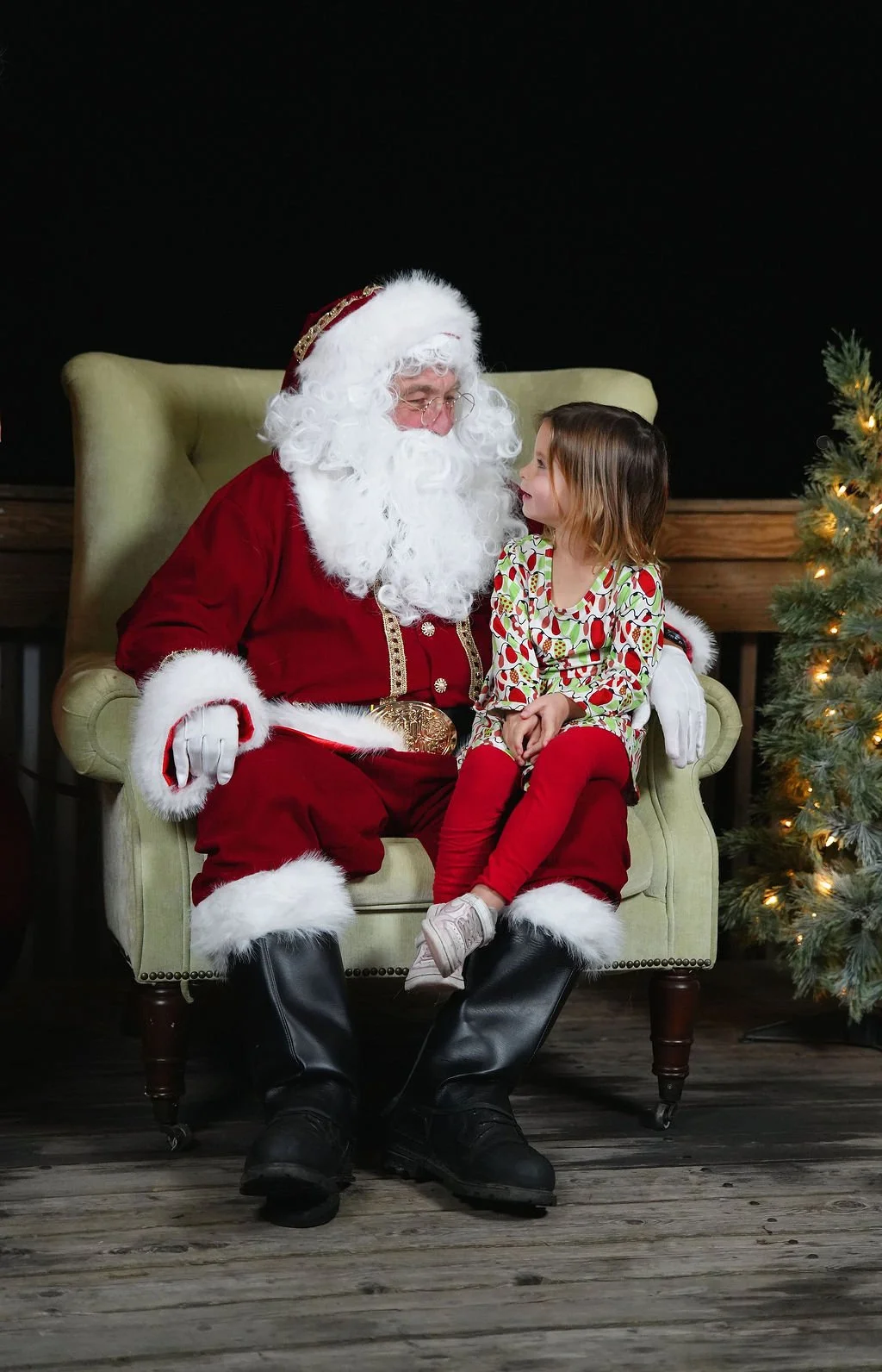 Santa Claus sitting on a green armchair with a young girl sitting on his lap, Christmas tree with lights on the right, dark background, and wooden floor.
