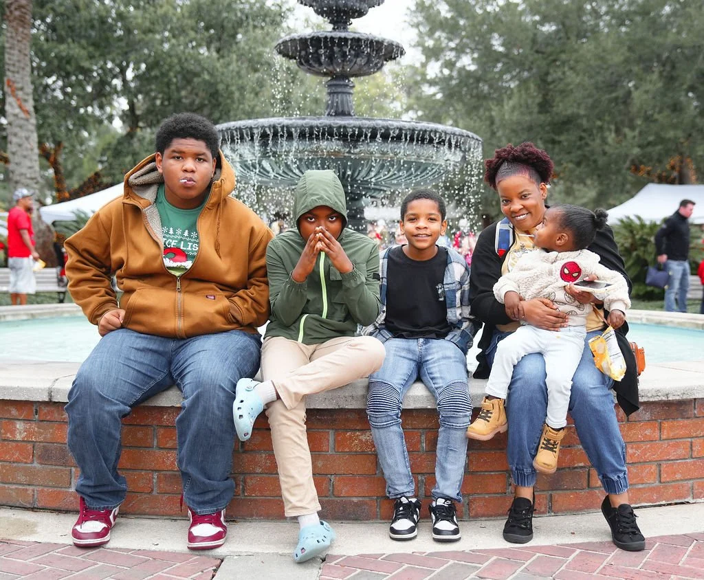 Five children sitting on a brick ledge in front of a fountain outdoors, with trees and tents in the background.