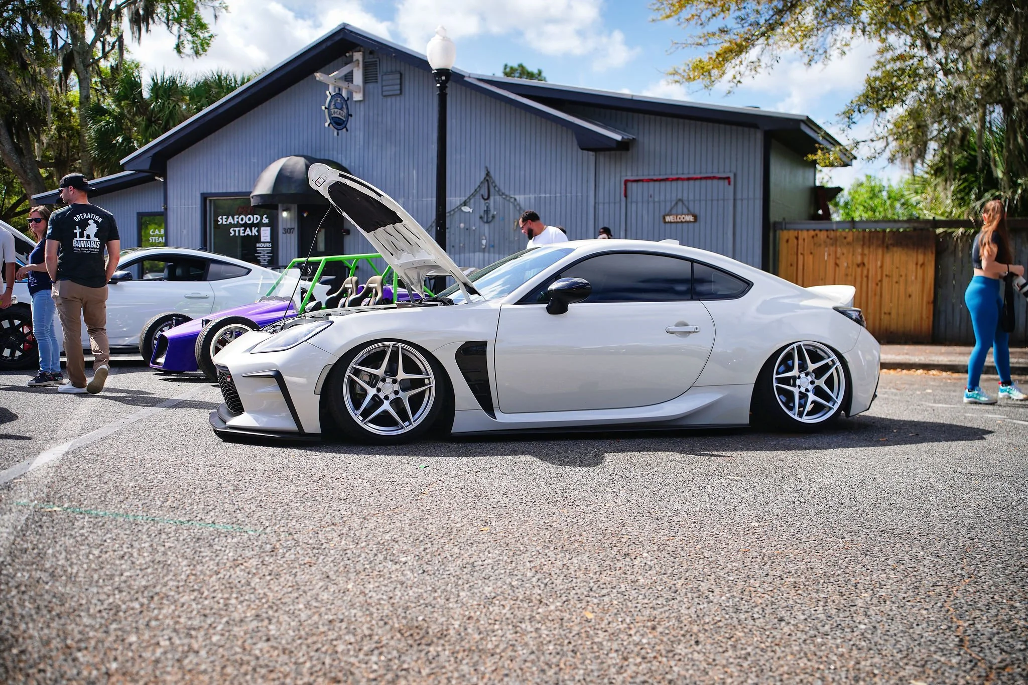 White sports car with lowered suspension and custom wheels parked at a car show, with other cars and people in the background.