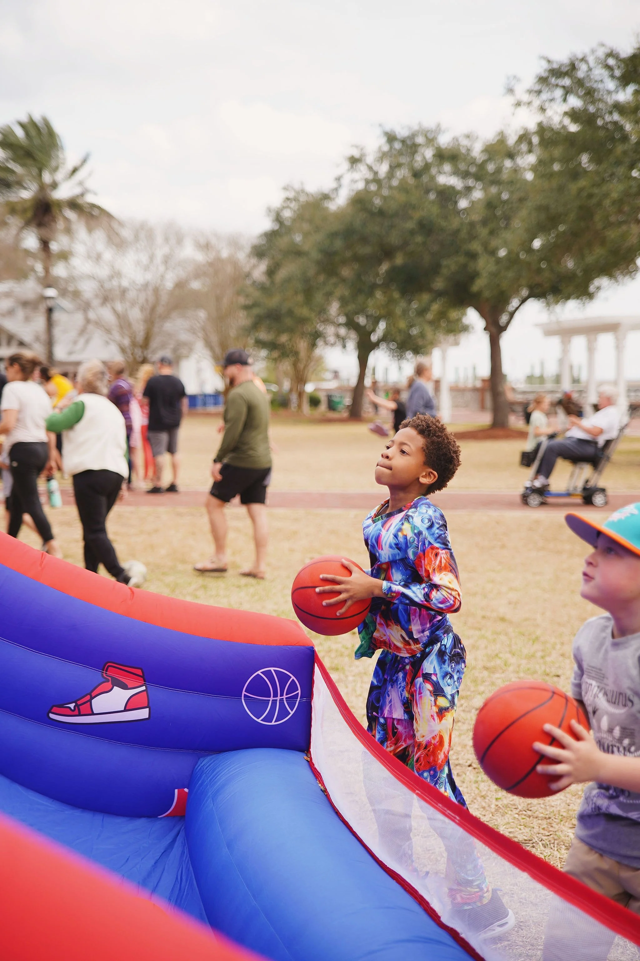 Two children holding basketballs near an inflatable basketball hoop at a park, with adults and trees in the background.