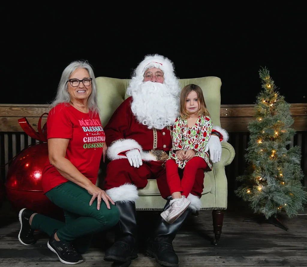 A woman, a man dressed as Santa Claus, and a young girl sitting on Santa's lap pose for a Christmas photo. The woman is kneeling on one knee, wearing glasses and a red Christmas shirt, smiling. The girl is wearing festive pajamas with snowmen and hol