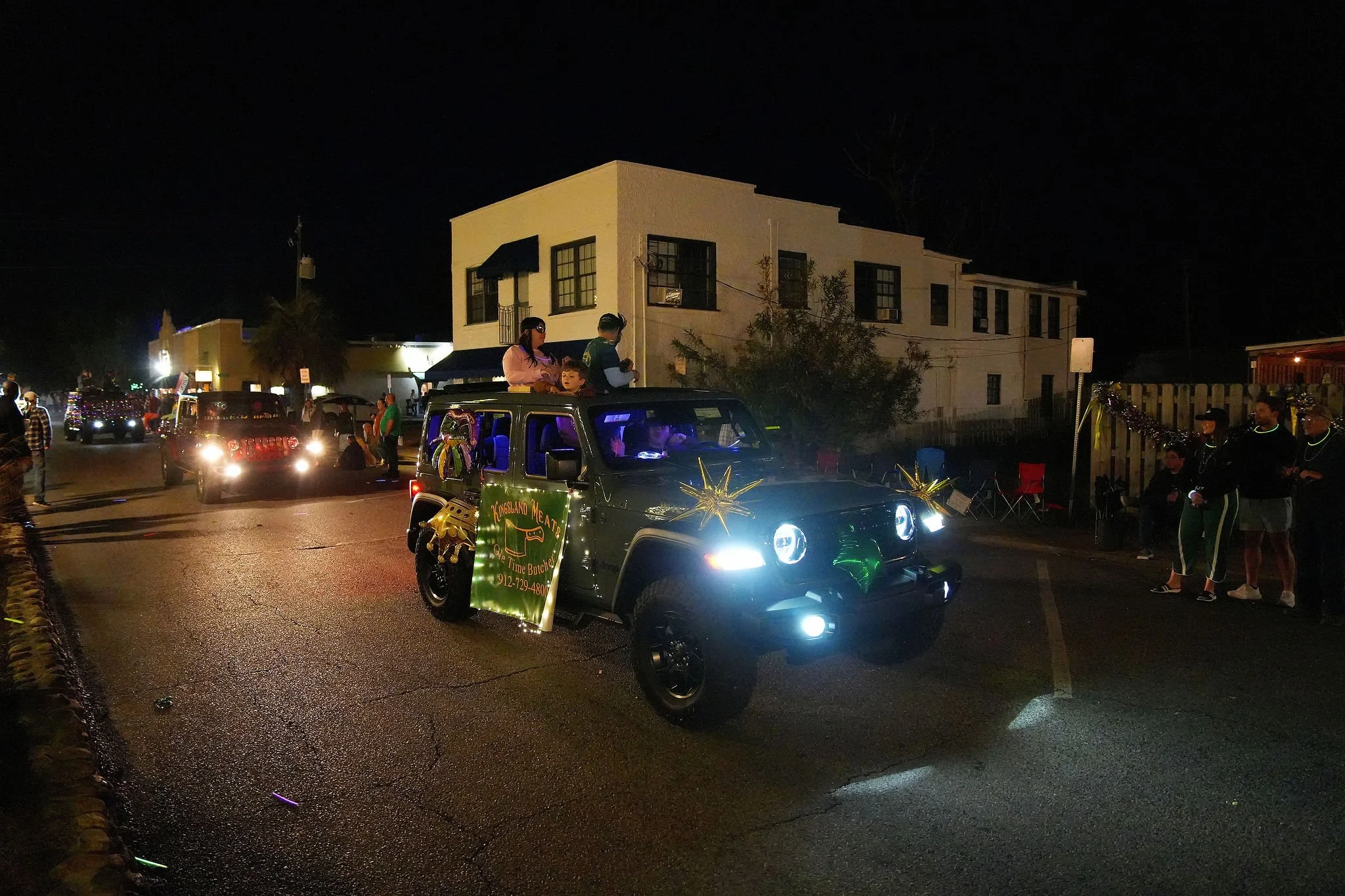 A nighttime parade featuring a decorated vehicle with people riding on top, surrounded by onlookers and other vehicles, illuminated by streetlights.