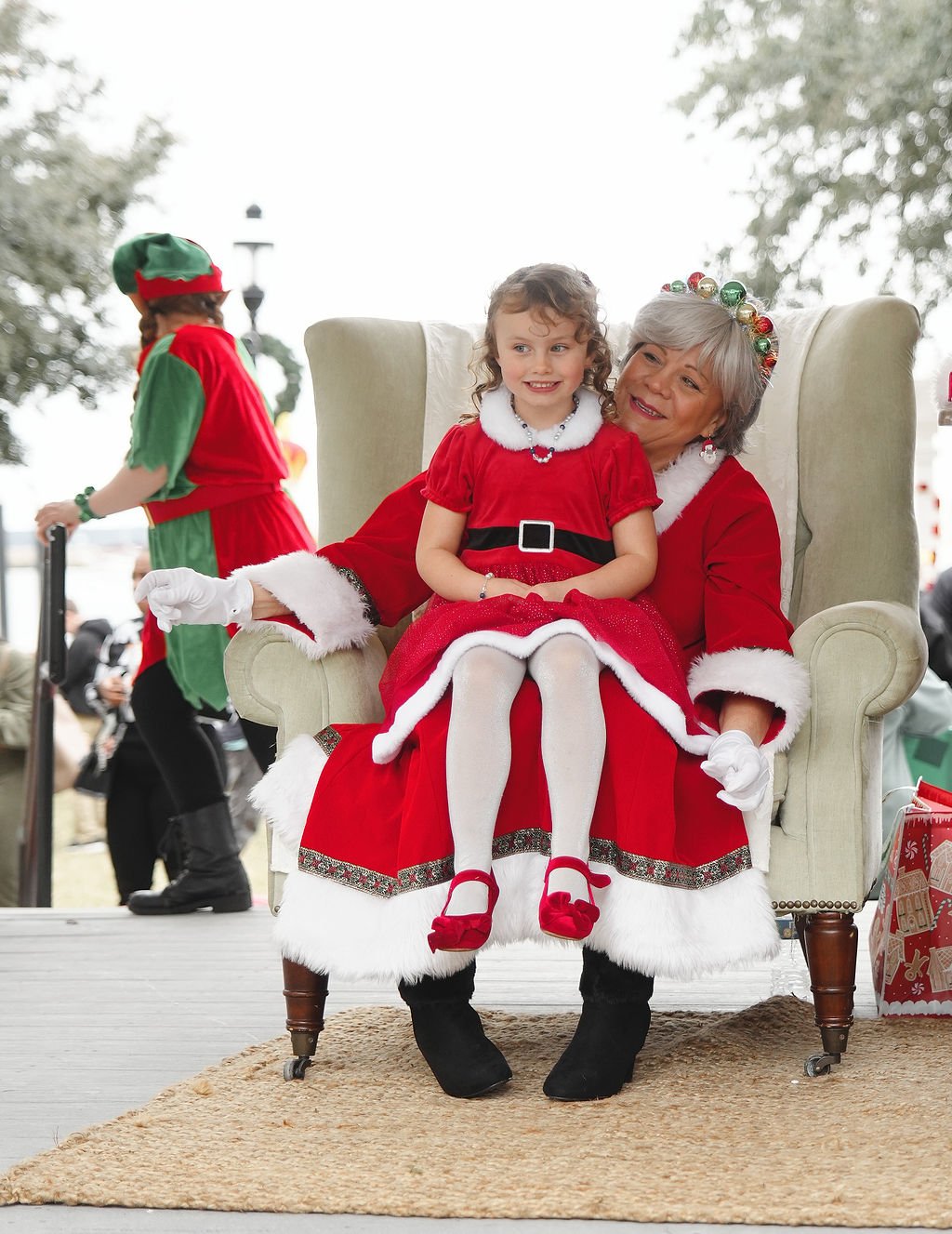 A young girl and older woman dressed as Santa Claus sitting together on a large, cream-colored armchair at a Christmas event. The girl is wearing a red dress with white trim and red shoes, and the woman is in a Santa costume with gray hair and festiv