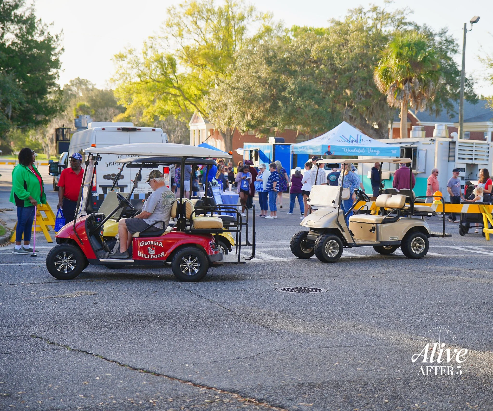 Two golf carts parked on a street at an outdoor event, with people walking around and vendor tents in the background. One cart has a Georgia Bulldogs logo, and there is a logo that says "Alive AFTER 5" in the bottom right corner.