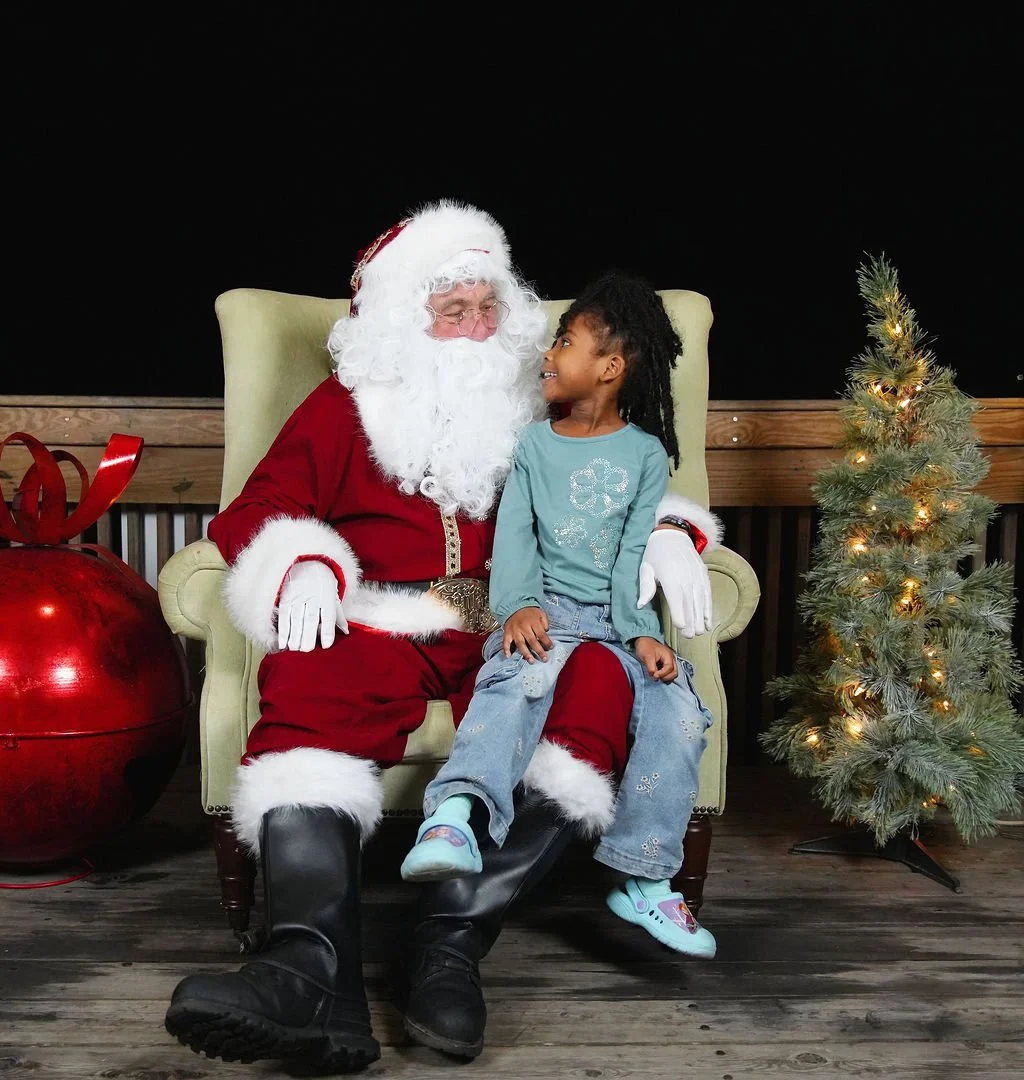 A young girl sitting on Santa Claus's lap, smiling and looking at him, with a decorated Christmas tree and a large red ornament in the background.