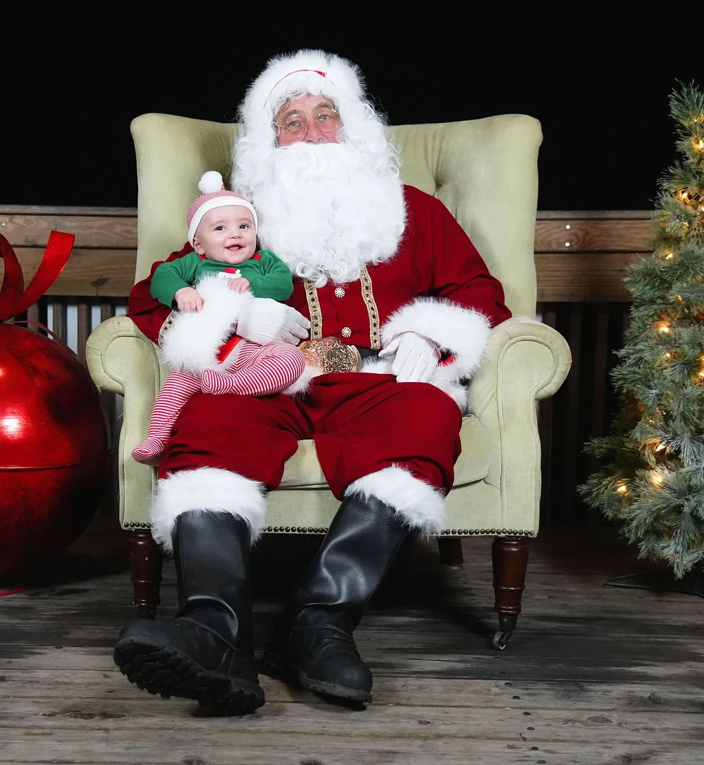 A young child in holiday clothing sitting on Santa Claus's lap, both smiling, with Christmas decorations including a decorated tree and large ornament in the background.