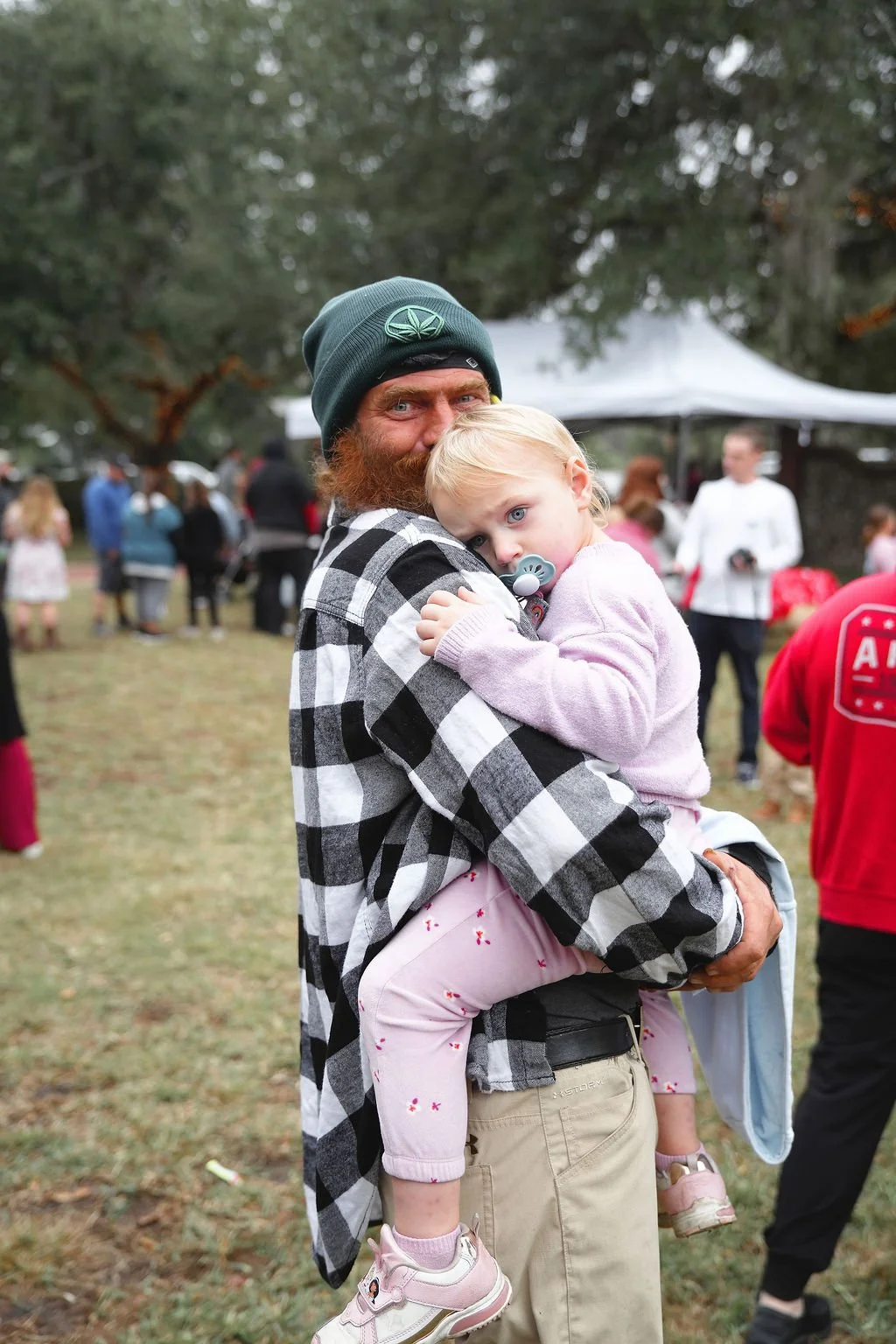 A man with a beard wearing a green beanie and a checkered shirt is holding a young girl with blonde hair, blue eyes, and a pacifier in her mouth. They are outdoors at a gathering with several people and trees in the background.