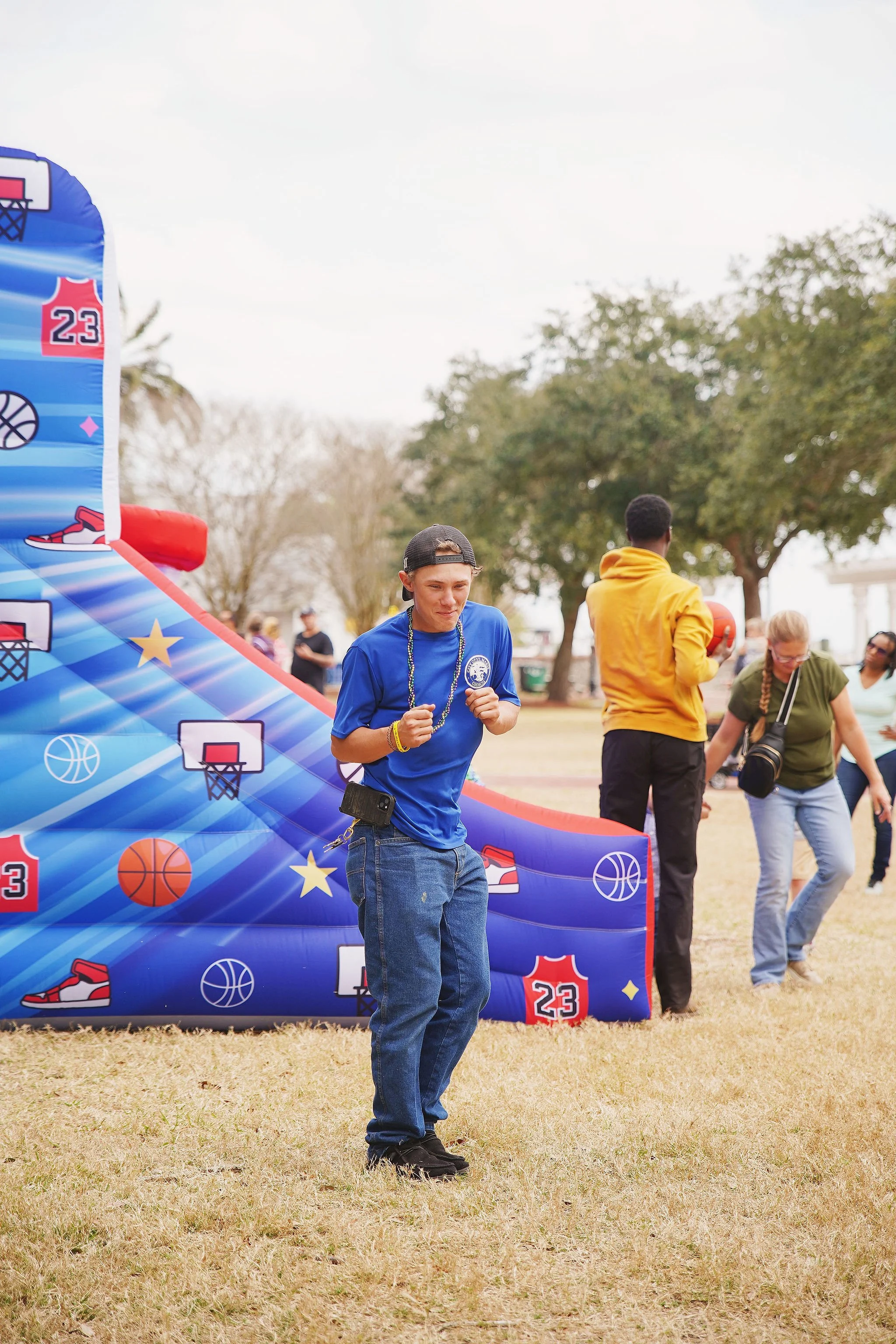 A young man in a blue shirt and baseball cap is dancing or jumping in front of an inflatable basketball game at an outdoor event, with other people in the background.
